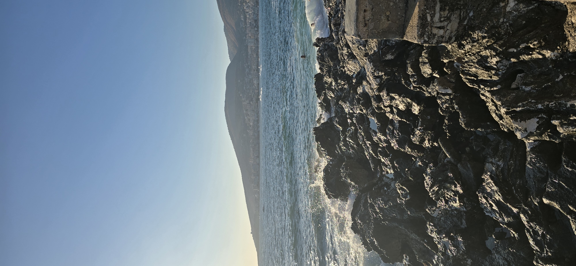 This image depicts a rocky coastline in Saranda, Albania, on a clear, sunny day. The foreground is dominated by large, dark, weathered rocks that are wet from the sea. Waves crash against these rocks, creating white foam and spray. In the middle ground, the blue sea extends towards the horizon. A person is visible in the water, swimming. Further in the background, a coastal city is nestled against a green, hilly landscape under a bright blue sky. The city appears to be Saranda, with numerous buildings and structures visible along the coastline and up the hillsides. Several boats are present on the water. The overall scene conveys a sense of a beautiful, natural coastal environment meeting an urban landscape.