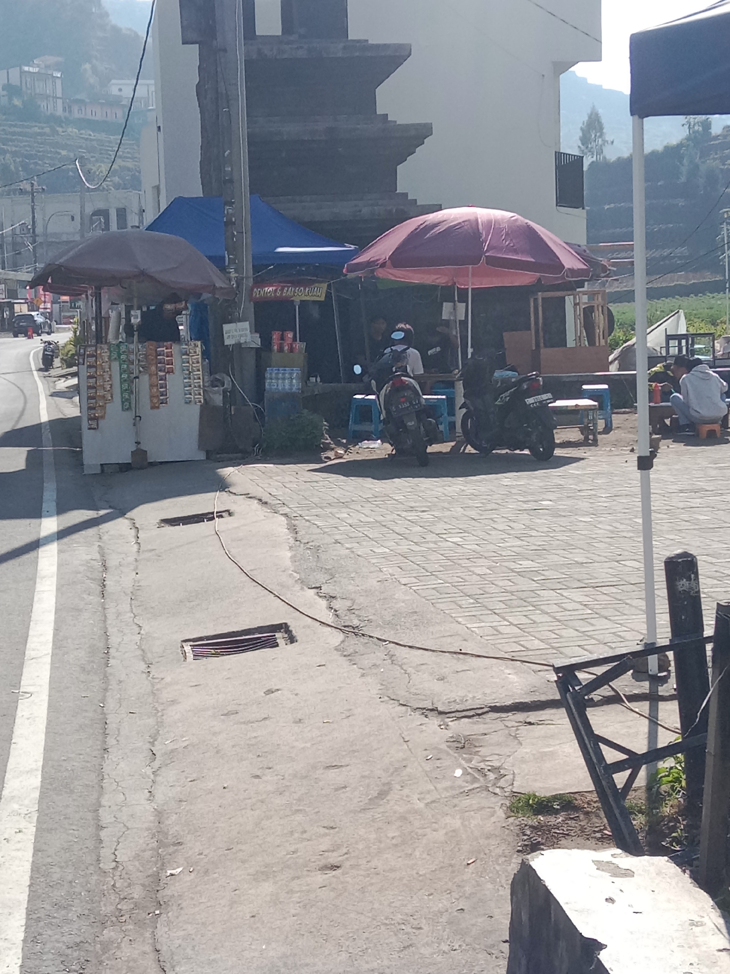 The image depicts a street scene in Parikesit, Indonesia, with several vendors and customers present. A man is seen serving food from a stall adorned with a dark red umbrella. Behind him, a blue tent is set up, also functioning as a vendor area. Two motorcycles are parked near the stalls. In the background, several buildings and terraced hills are visible under a bright sky. The foreground shows a paved walkway and a road with white lane markings.