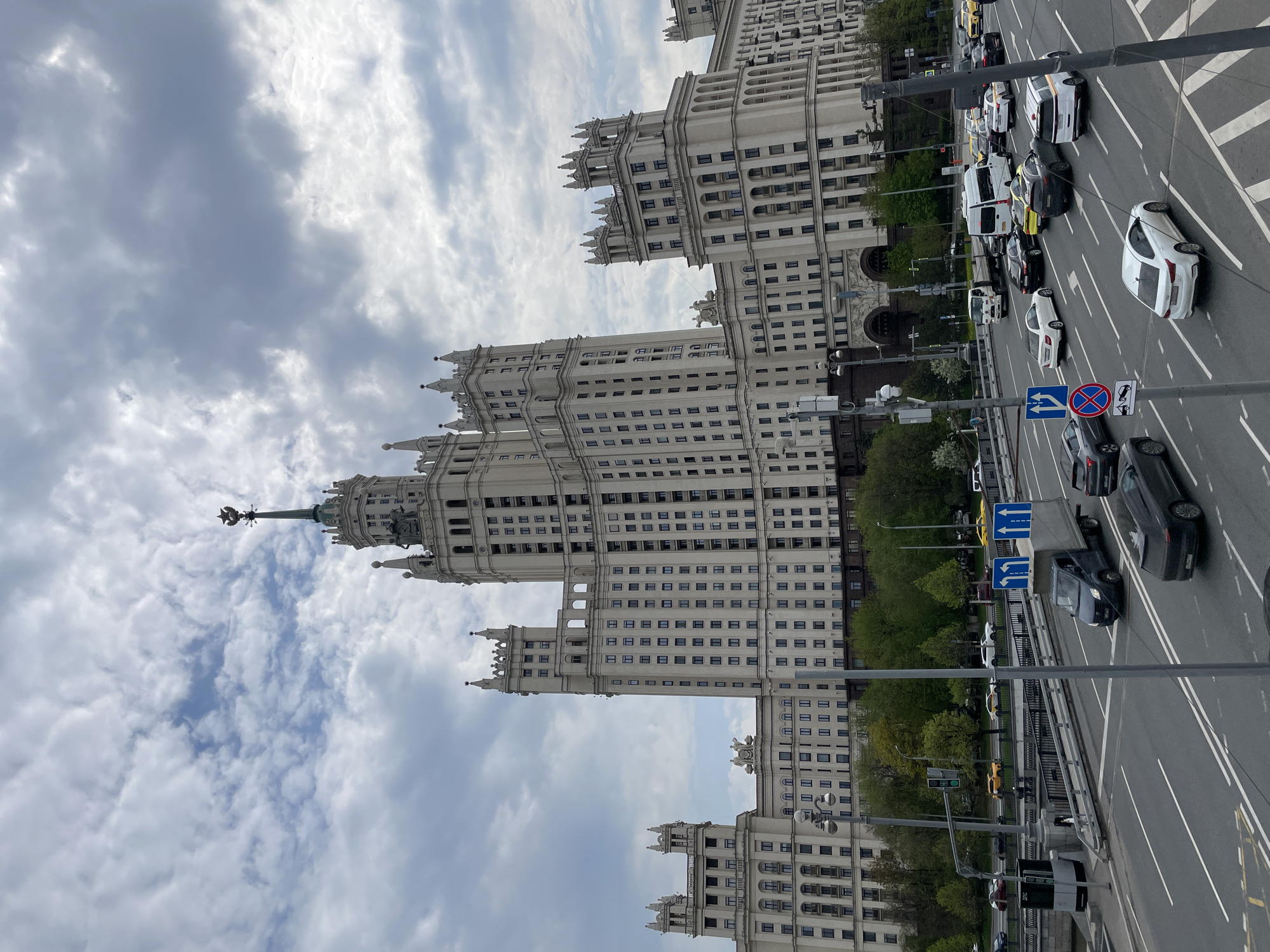The image shows the Kotelnicheskaya Embankment Building in Moscow, Russia, taken from an elevated position. The Stalinist-era skyscraper dominates the view, set against a cloudy sky. Below the building, a wide road carries traffic. Cars, vans, and buses are visible on the roadway. Street signs are also present, indicating traffic directions. Trees line parts of the road, adding greenery to the urban environment.