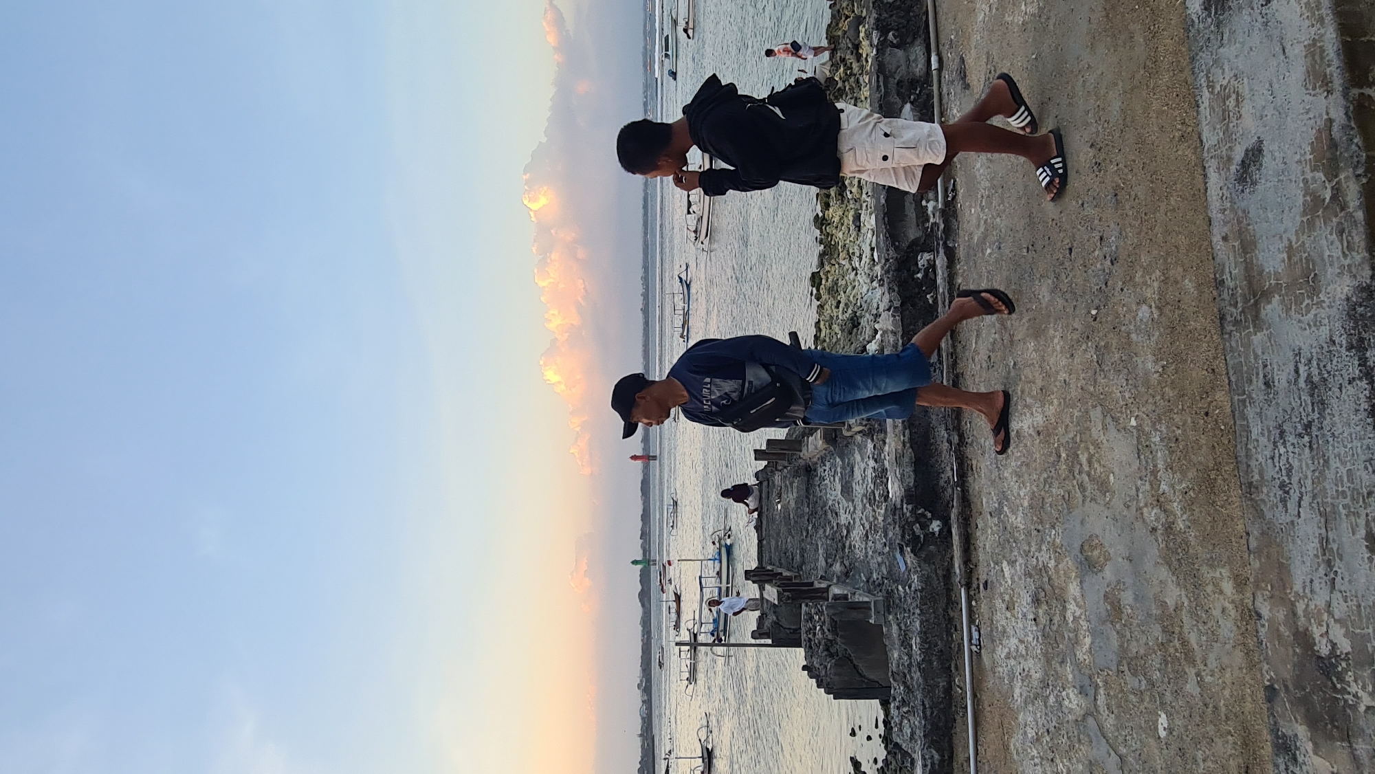 The image captures a serene coastal scene in Denpasar, Indonesia, likely during the golden hour of sunset, given the warm hues illuminating the sky and clouds. Two individuals are the primary subjects, standing on a weathered concrete pier or seawall.
The person in the foreground, wearing a blue long-sleeved shirt, blue shorts, and a black baseball cap, is looking down towards the water. A black cross-body bag is slung over their shoulder. They are wearing dark flip-flops.
Slightly behind and above this individual is another person dressed in a black hoodie and white cargo shorts. This person is also looking downwards, possibly interacting with something or the other person. They are wearing black flip-flops with white stripes.
The background reveals a calm body of water dotted with numerous small boats, some appearing to be traditional Indonesian fishing vessels. The water's surface reflects the soft light. In the distance, a few more people can be seen along the pier or shore. The sky is a gradient of soft blue transitioning to warm orange and yellow tones, indicative of sunset. The overall atmosphere seems peaceful and relaxed.