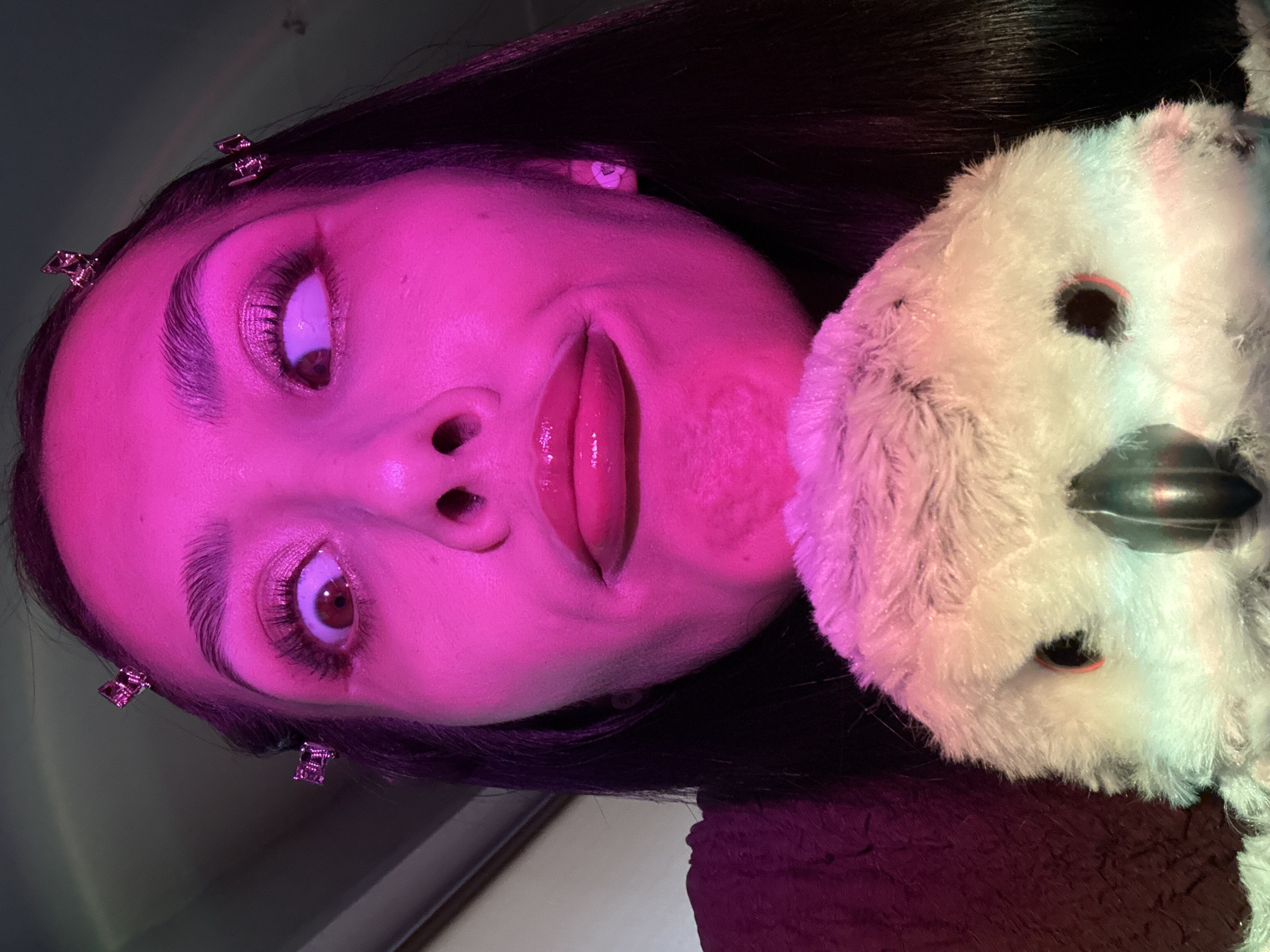 A selfie-style image captures a person with dark hair, illuminated by pink lighting. The individual's face is prominently displayed, showing features like eyes with visible lashes, eyebrows, nose, and glossy lips. Hair clips are visible on the side of the head. Adjacent to the person's face is a white, plush toy with a black beak and beady eyes. The environment and specific location in Votkinsk, Russia, cannot be determined from this image alone.