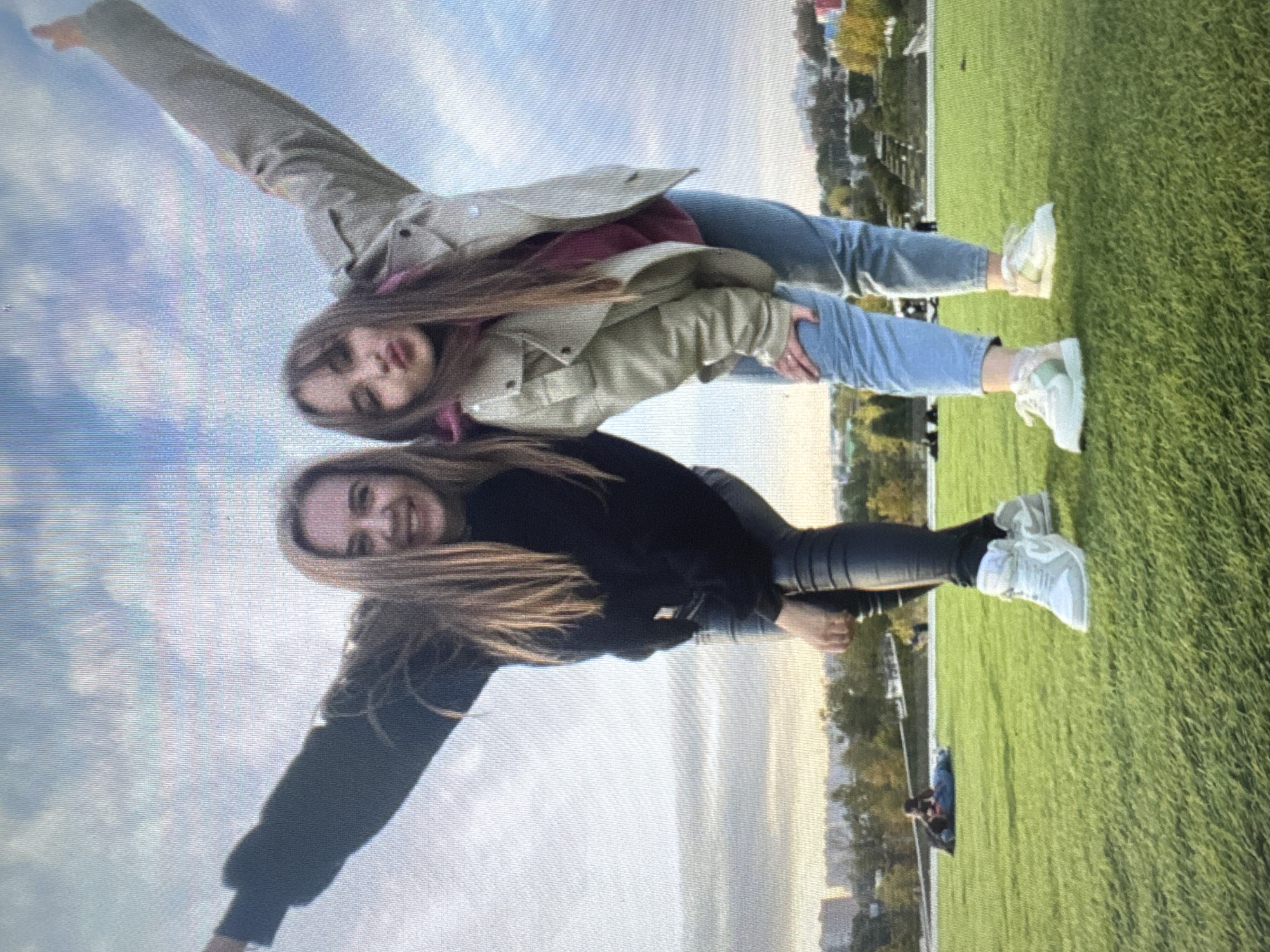 Two young women are standing in a grassy park. The woman on the left is wearing a black long-sleeved top and black leggings. She has her arms outstretched, with her left arm raised above her head. The woman on the right is wearing a beige jacket over a burgundy hoodie and blue jeans. She has her right arm raised above her head, and her left hand resting on her left knee. Both women are smiling at the camera. In the background, there are trees, buildings, and a cloudy sky. Two people are lying on the grass in the distance.