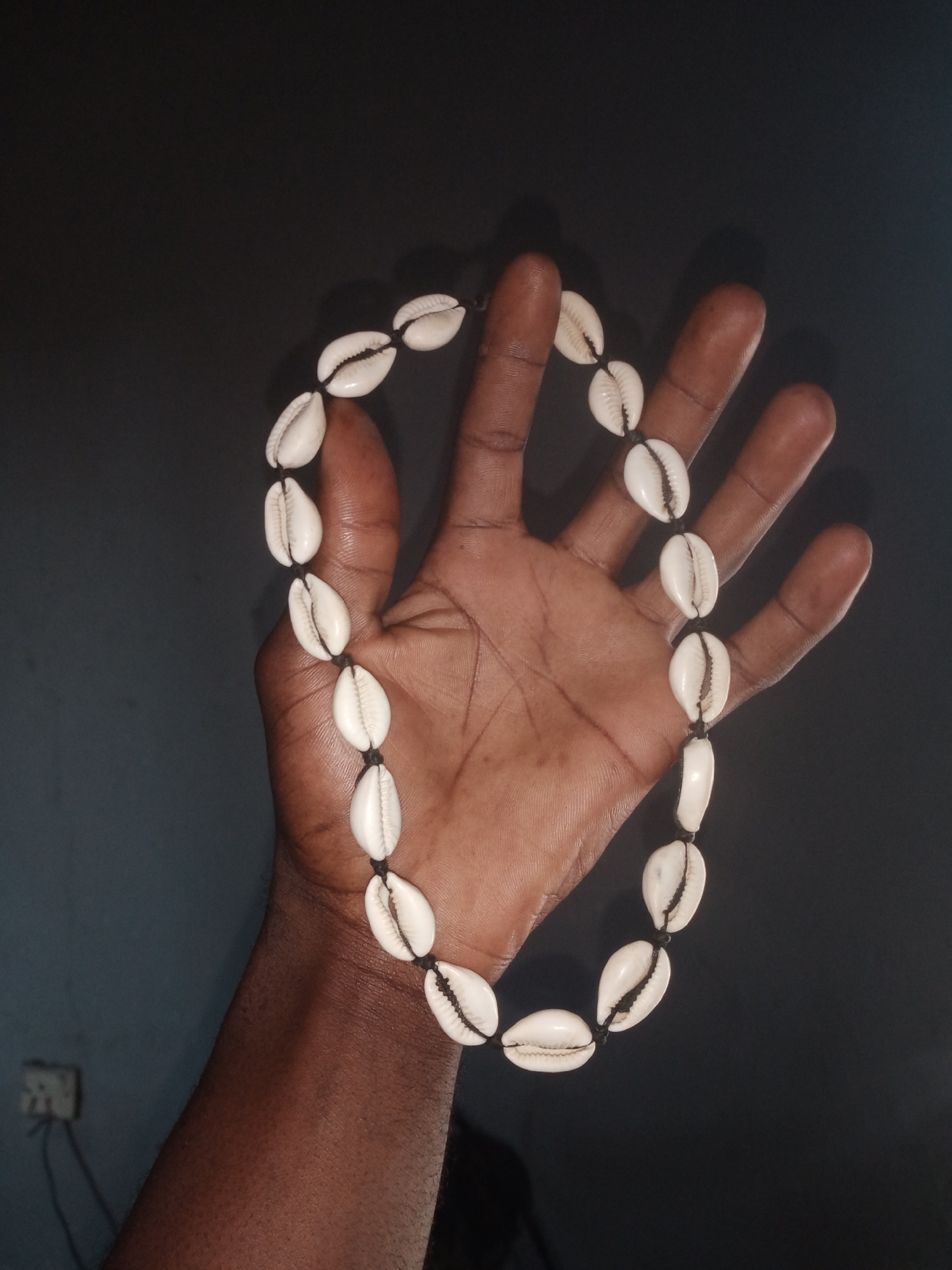The image shows a person's hand, with dark skin tone, holding a necklace made of cowrie shells. The necklace is black cord strung with numerous white cowrie shells, forming a circular shape across the palm and fingers. The background is a dark, plain wall. In the lower left corner of the image, a white electrical outlet with visible wires is mounted on the wall. The overall lighting is dim, casting soft shadows. The scene appears to be indoors. It is not possible to determine the specific location within Jalingo, Nigeria, based on the visual information provided.