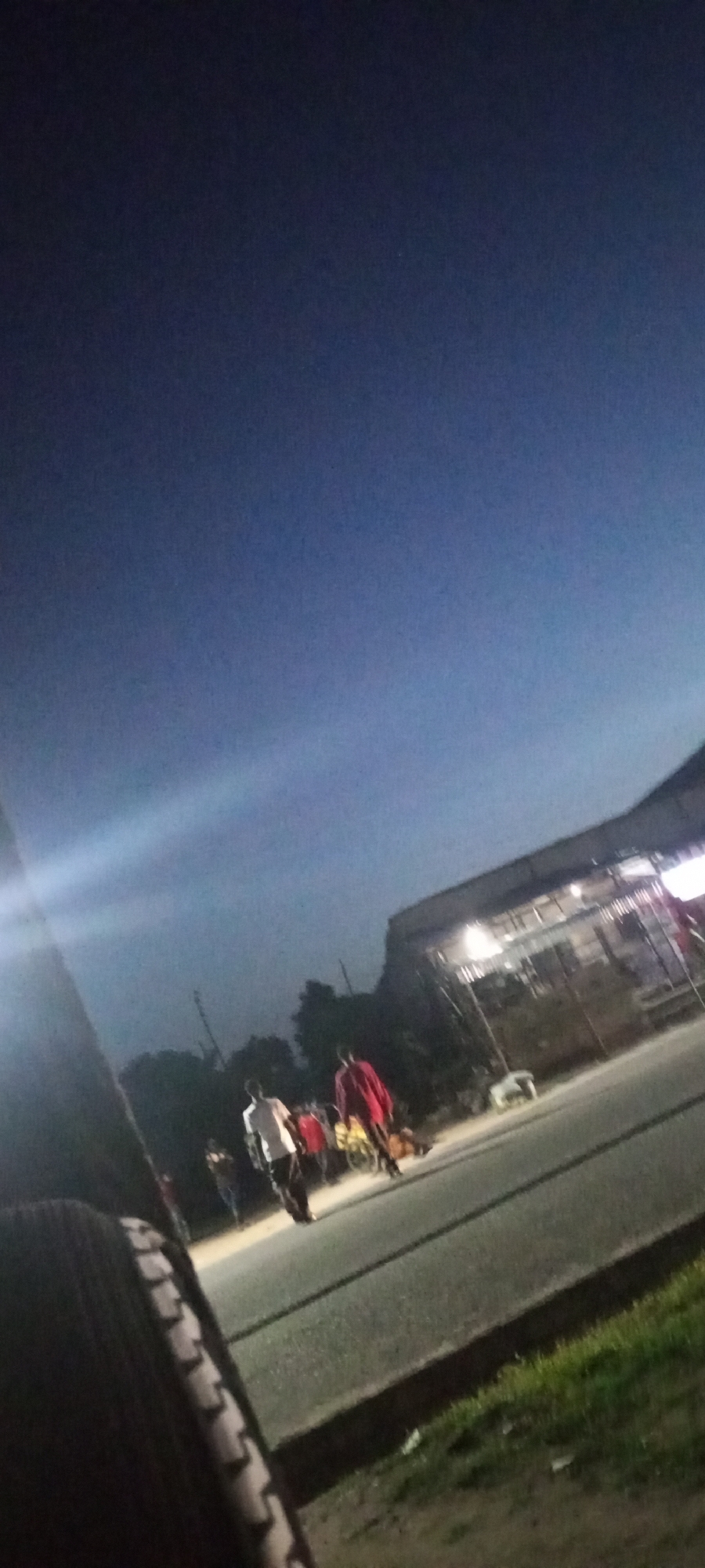 The image depicts a twilight scene on a paved road in Jalingo, Nigeria. In the foreground, the tread of a tire is visible on the left side. A group of people are gathered on the side of the road, with some individuals walking and others standing. One person in the foreground is wearing a white shirt and dark pants, while another to their right is wearing a red shirt and dark pants. In the background, there appears to be a structure with a sloped roof, illuminated by artificial lights. Trees are visible behind the people, and the sky is a deep blue, indicating the transition from day to night. The overall impression is of a casual gathering or activity taking place outdoors in a semi-urban environment.