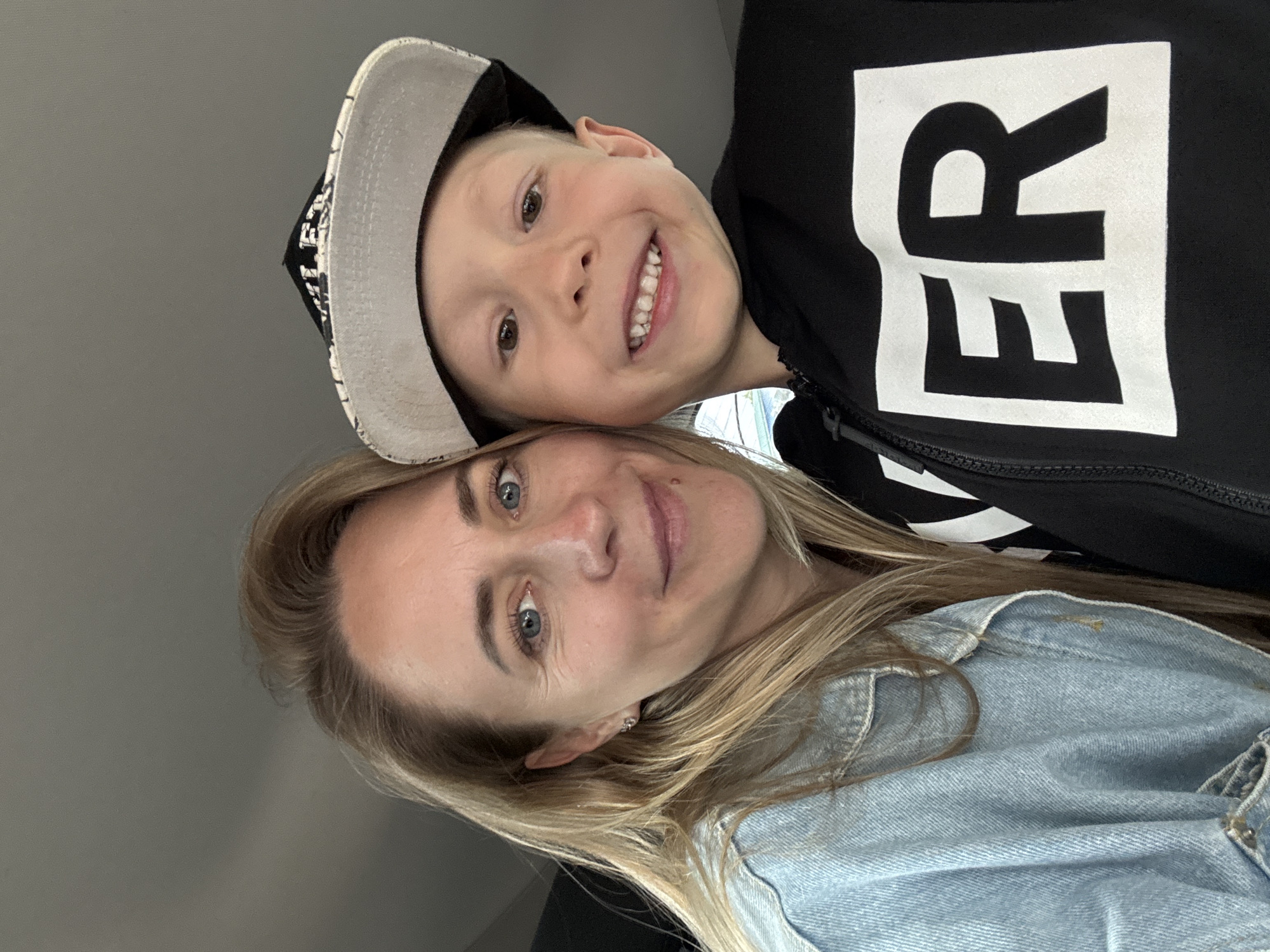 In a close-up, self-taken photograph, a woman with blonde hair and blue eyes, wearing a light blue denim jacket, is positioned on the left side of the frame. To her right, a young boy with blonde hair and brown eyes is wearing a black hoodie with large white lettering that spells out "ER" within a white rectangle. He is also wearing a black and gray flat-brimmed baseball cap. Both individuals are smiling and looking towards the camera. The background is indistinct, appearing to be an indoor setting with a muted gray ceiling. The image does not provide any visual cues to suggest a location within the city of Chaikovsky, Russia.