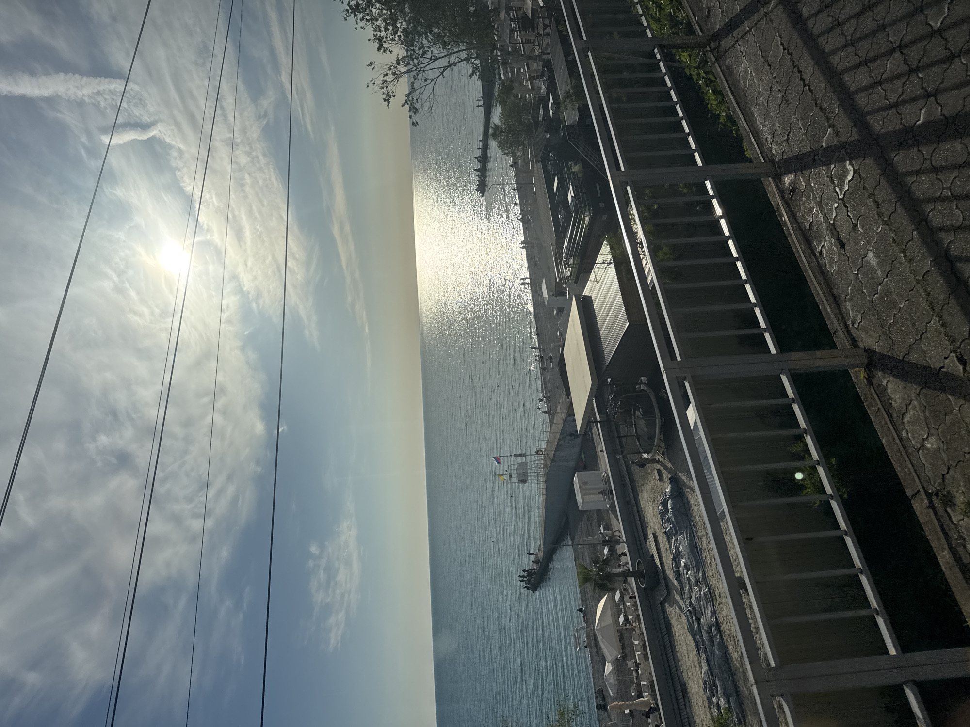 The image displays a sunny coastal scene in Sochi, Russia, captured during the daytime, likely late morning or afternoon. A vast body of water, presumably the Black Sea, dominates the center of the frame, shimmering intensely from the direct reflection of the sun, which is visible towards the upper left. The sky above is bright, with scattered wispy white clouds and several thin, dark lines, possibly power cables, stretching across it.
To the right, a paved promenade or walkway runs along the shoreline, bordered by a prominent metal railing. Along this coast, a pier extends into the water, with several small figures, likely people, visible on it. A white lifeguard stand with a red and white roof is noticeable near the pier, and a small Russian flag (white, blue, red tricolor) can be seen flying from a pole, subtly confirming the location as Sochi. Other elements of a beach or resort environment, such as umbrellas, lounge chairs, and parts of buildings or cafes, are also present along the water's edge, interspersed with trees and greenery.
Strong shadows from an unseen structure fall across the paved ground and the metal railing in the lower right foreground, indicating the sun is at an angle. The overall impression is one of a bright, leisurely day by the sea. No discernible text is visible in the image.