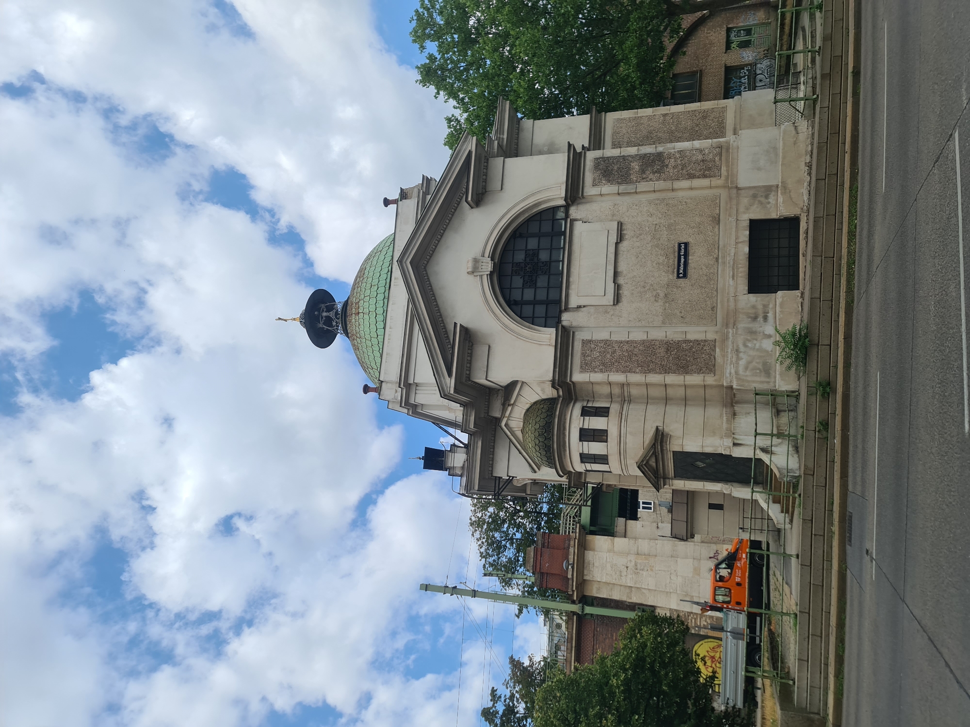 This is a medium-shot, outdoor, eye-level photograph of the St. Rochus Cemetery's Columbarium in Vienna, Austria. The Columbarium is a cream-colored, classical building with a green dome topped by a small, dark cupola with a golden cross. The building features a large arched window with decorative metalwork on the facade. To the left of the Columbarium is a tall, concrete structure with red brick accents and overhead power lines. A bright orange utility truck is parked on the left side of the image, near a set of stairs leading up to the building. The ground in front of the building is paved with gray concrete slabs and bordered by a green metal fence. Trees with lush green foliage are visible behind and to the right of the Columbarium. The sky is a bright blue with scattered white clouds. A street with white lane markings is in the foreground. The street sign visible on the building reads "9. Währinger Gürtel."
