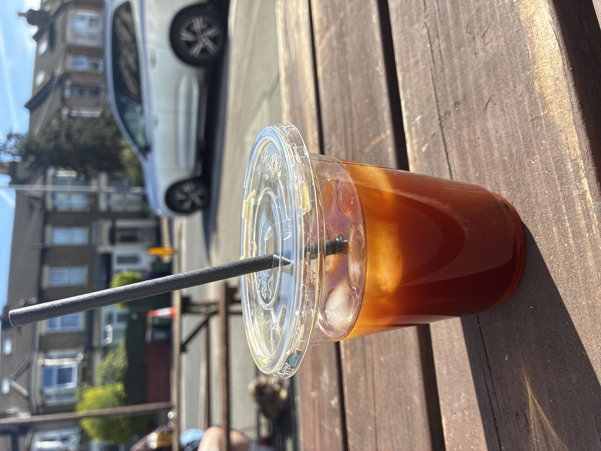 The image shows a plastic cup containing a beverage with ice, resting on a wooden surface. A black straw protrudes from the cup's lid. In the background, blurred buildings, trees, and a white car are visible. The scene appears to be outdoors during daylight, with strong sunlight casting shadows on the wooden surface. The specific location in the city of London, United Kingdom cannot be determined from the image alone.