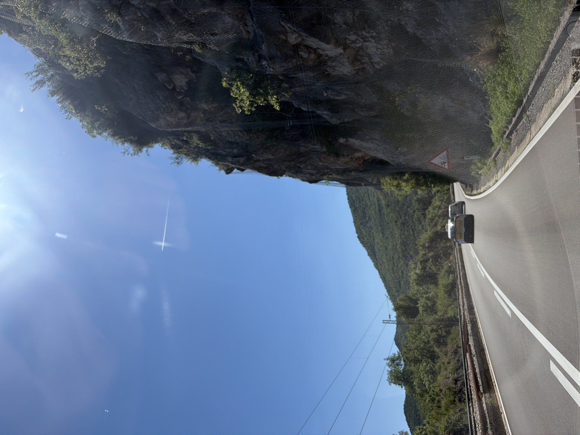 The image depicts a highway in Kruševica, Montenegro, under a clear blue sky with bright sunlight. A winding road with white lane markings is visible, with two cars traveling ahead. To the right, a steep, rocky cliff face is protected by a mesh netting system. Vegetation, including trees and shrubs, grows on the cliff and the surrounding hillsides. A railway track runs parallel to the road on the left. A triangular warning sign with a slippery road symbol is posted near the cliff.