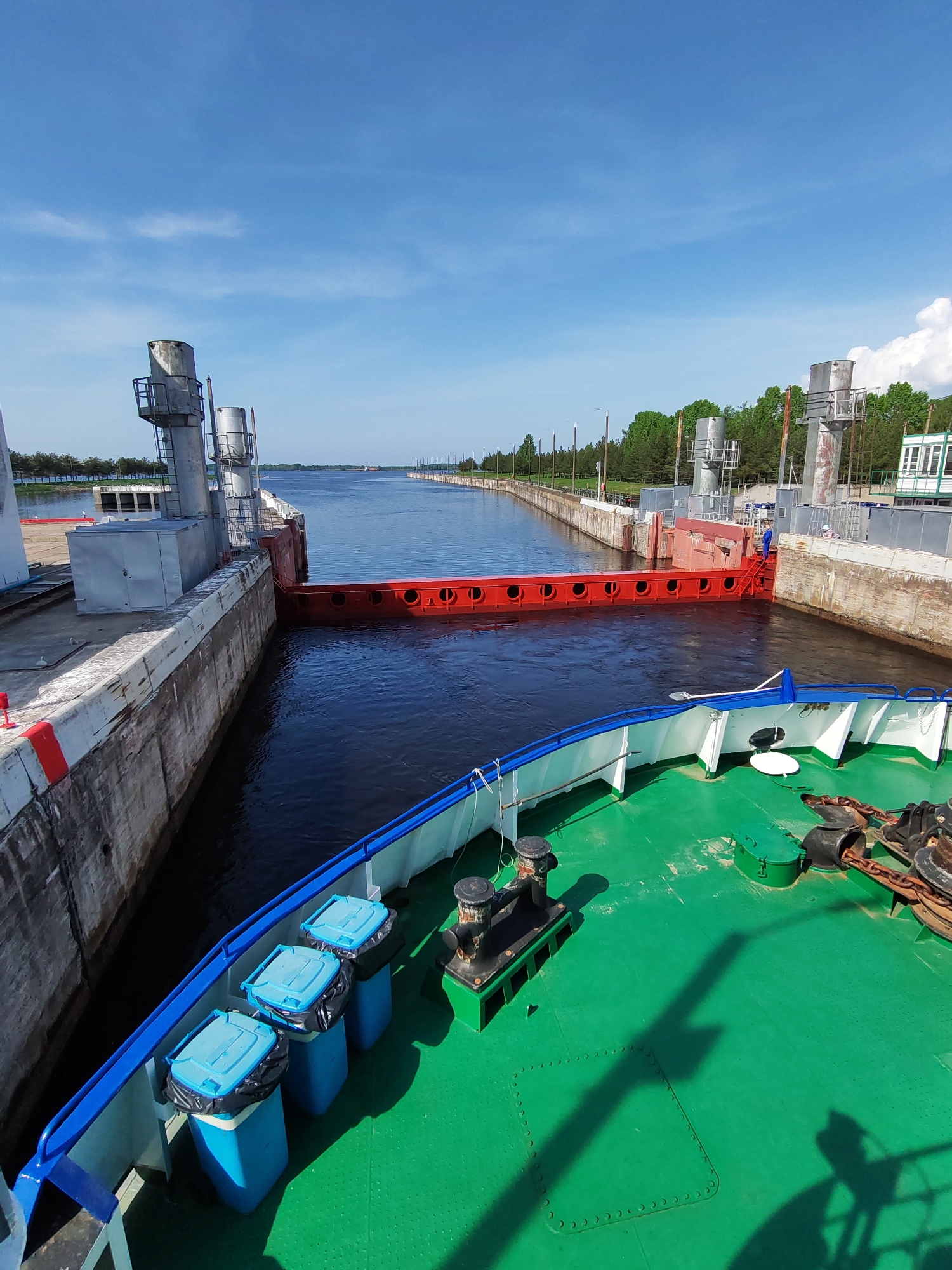 The image depicts a view from the deck of a ship, likely a river vessel, positioned within a canal lock. The lock is closed, indicated by a red gate spanning the width of the canal. The surrounding walls of the lock are concrete. On the left side of the canal are metal structures and a low building. The canal stretches out into the distance, bordered by trees and utility poles. The sky is clear and blue with some clouds. On the ship's deck in the foreground, there are blue containers, mooring bollards, and anchor chains. The canal lock is located in Sheksna, Russia. A man is standing on a red ladder near the gate.
