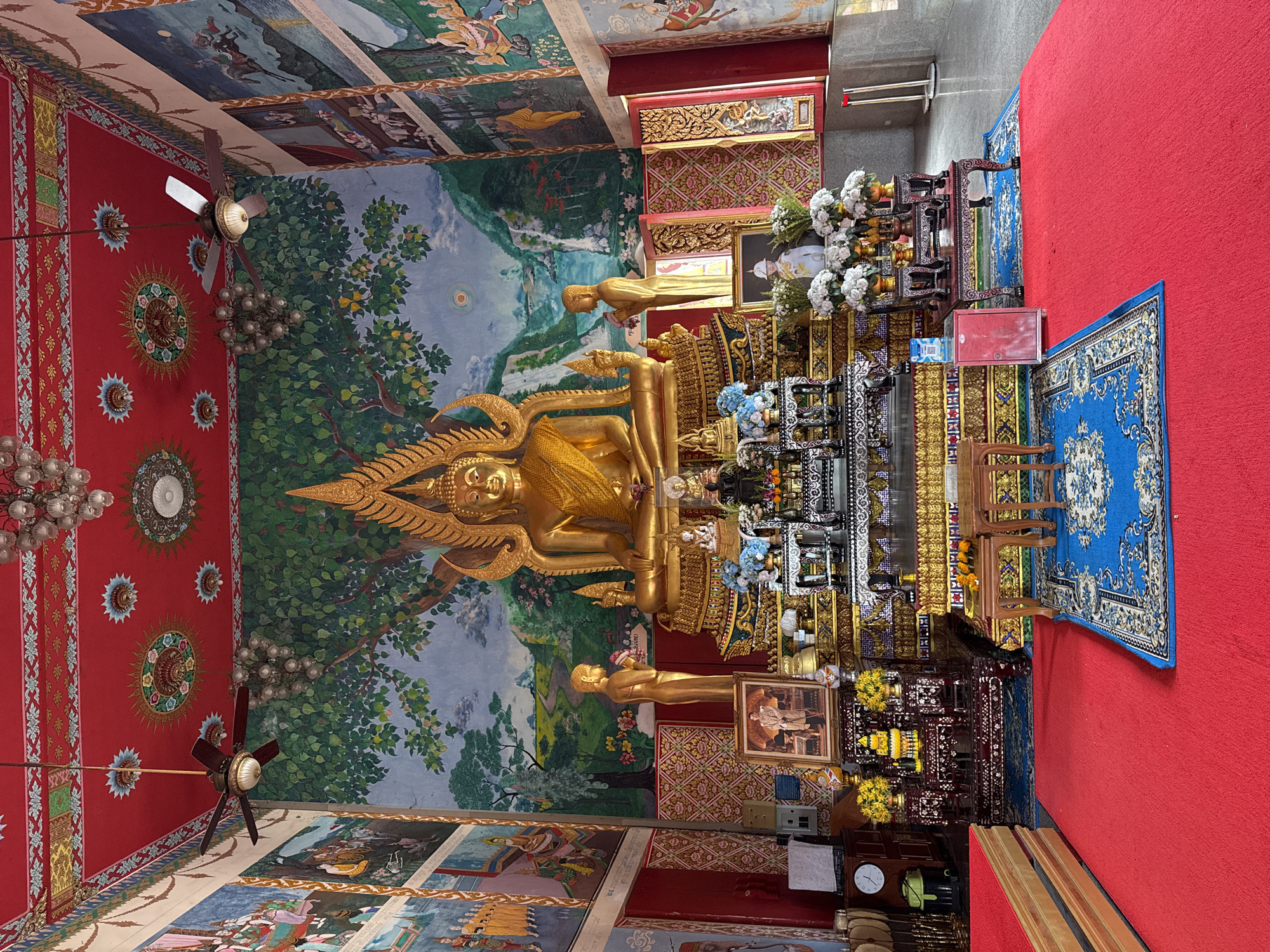 This image depicts the interior of a Buddhist temple in Ko Samui District, Thailand. The central focus is a large, ornate golden Buddha statue seated on a multi-tiered altar. The altar is adorned with intricate carvings, offerings, and floral arrangements. Flanking the main statue are two smaller golden figures. The walls of the temple are covered with vibrant murals depicting scenes from Buddhist narratives and landscapes. The ceiling is painted red and decorated with intricate patterns and ornate light fixtures, with two ceiling fans visible. The floor is covered with red carpeting, with a blue rug placed in front of the altar. Several small wooden tables and a red cabinet are also present in the foreground. The overall atmosphere is one of reverence and cultural richness.