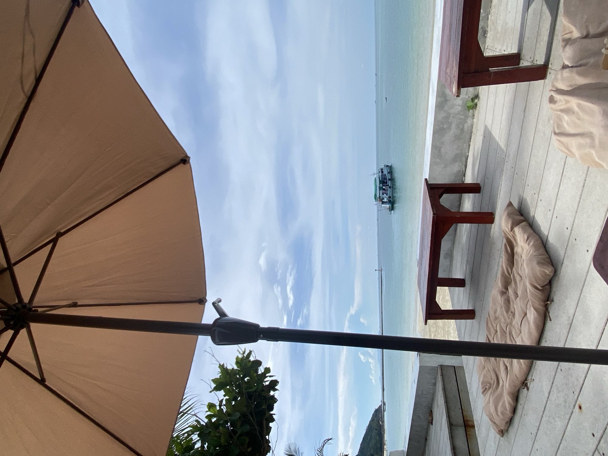 View from an outdoor deck at Baan Chaloklum, Thailand. The deck has wooden benches and cushions. A large tan umbrella shields the seating area from the sun. Beyond the deck, the ocean extends to the horizon, with a boat in the water. The sky is blue with scattered clouds. Green foliage grows below the deck.