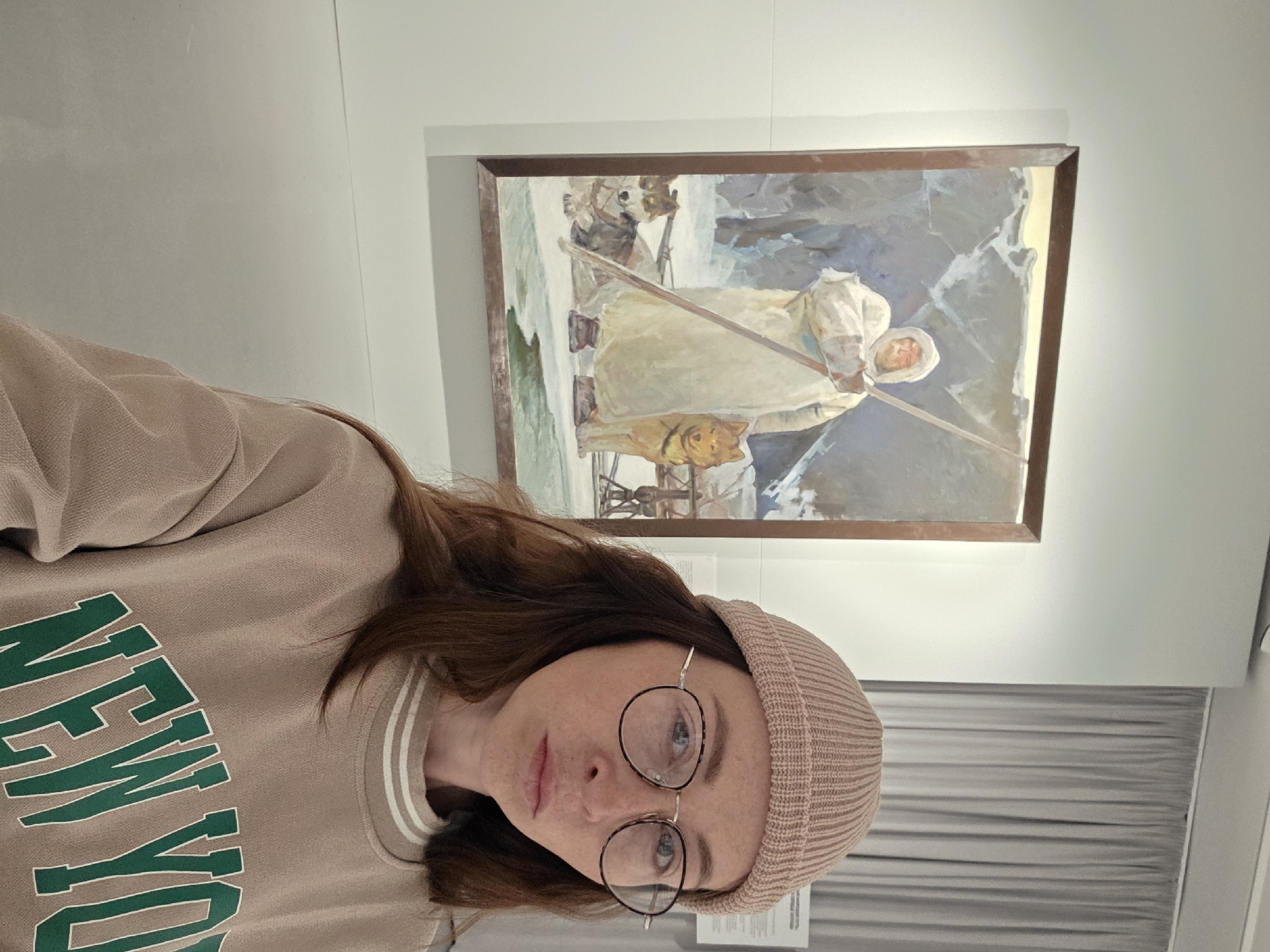 The photograph appears to be a selfie taken inside a gallery or museum in the city of Chaikovsky, Russia. A person with brown hair, wearing a beige sweatshirt with "NEW YORK" written in green letters, a knitted cap, and round eyeglasses is posing in the foreground. Behind the person is a framed painting depicting a woman in a white garment, accompanied by dogs and what appears to be a sled, set against a mountainous, snowy landscape. The background also includes what seems to be a wall or room divider with horizontal lines, possibly curtains or blinds. The lighting suggests an indoor setting.