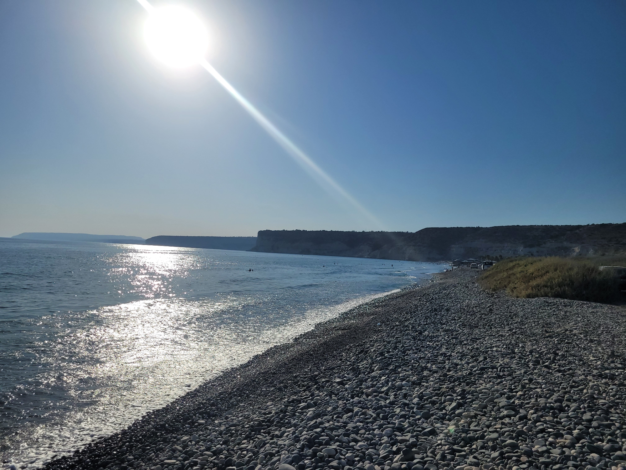 The image depicts a wide, pebbled beach in Episkopi, Cyprus, under a bright, sunny sky with the sun positioned in the upper left quadrant, casting a strong glare on the scene. The sea is a calm, blue expanse, reflecting the sunlight with numerous sparkles. Several people are visible in the water, some swimming and others wading. The coastline stretches into the distance, with a rocky cliff face visible on the horizon. The beach itself is covered in smooth, grey pebbles of varying sizes, and sparse vegetation grows along the edge of the cliff. In the foreground, the pebbled beach dominates the right side of the frame, sloping gently towards the water.