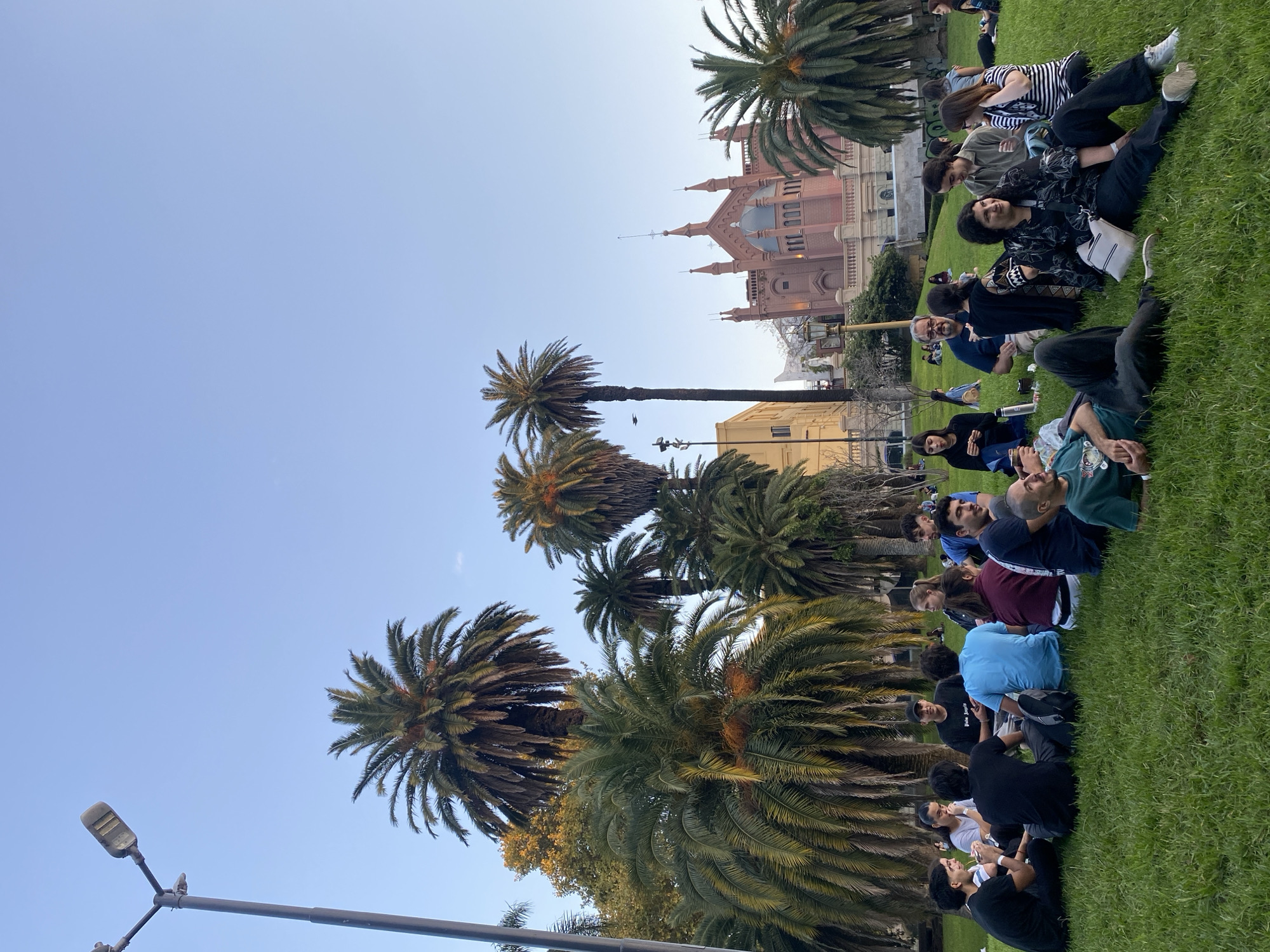 An outdoor scene captures a diverse group of people gathered on a lush green lawn under a clear blue sky. The image is taken from a slightly low angle, emphasizing the tall palm trees that dominate parts of the frame.
In the foreground and mid-ground, individuals, mostly young adults, are seated casually on the grass, forming several informal groups and lines. They are dressed in casual attire, including t-shirts, shirts, and trousers. Many appear to be engaged in conversation or listening intently, with relaxed and attentive expressions, suggesting a social gathering, a class, or an outdoor meeting. One person is wearing a black t-shirt with the words "Palm Angels" visible on it. A thermos bottle is also seen near one of the groups.
Behind the gathering, a striking architectural landmark stands out: a large, ornate building constructed from red brick, featuring multiple spires and intricate details. This structure suggests a historic institution or a religious building. Other less distinct buildings are also visible further in the background. Numerous tall palm trees, with both thick trunks and dense green canopies, are scattered throughout the park, contributing to a warm, subtropical atmosphere. A streetlamp post is visible in the lower left portion of the image.
The clear sky and soft, even lighting suggest pleasant weather, likely late afternoon or early evening. The setting appears to be a public park or garden adjacent to an urban area, consistent with the provided location context of Buenos Aires, Argentina.
