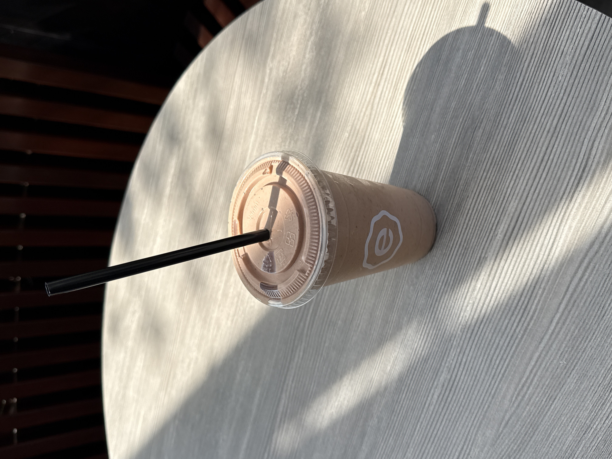 The photograph shows a chocolate milkshake on a light grey round table in West Hollywood, California, USA. The milkshake is in a clear plastic cup with a translucent brown lid and a black straw. The side of the cup has a white logo featuring a stylized letter "e". The table surface has a subtle linear pattern. The lighting suggests a sunny day with a distinct shadow cast on the table, possibly from a nearby tree or structure. Some dark vertical bars can be seen in the background, likely part of an outdoor seating arrangement. No people are visible in the frame.