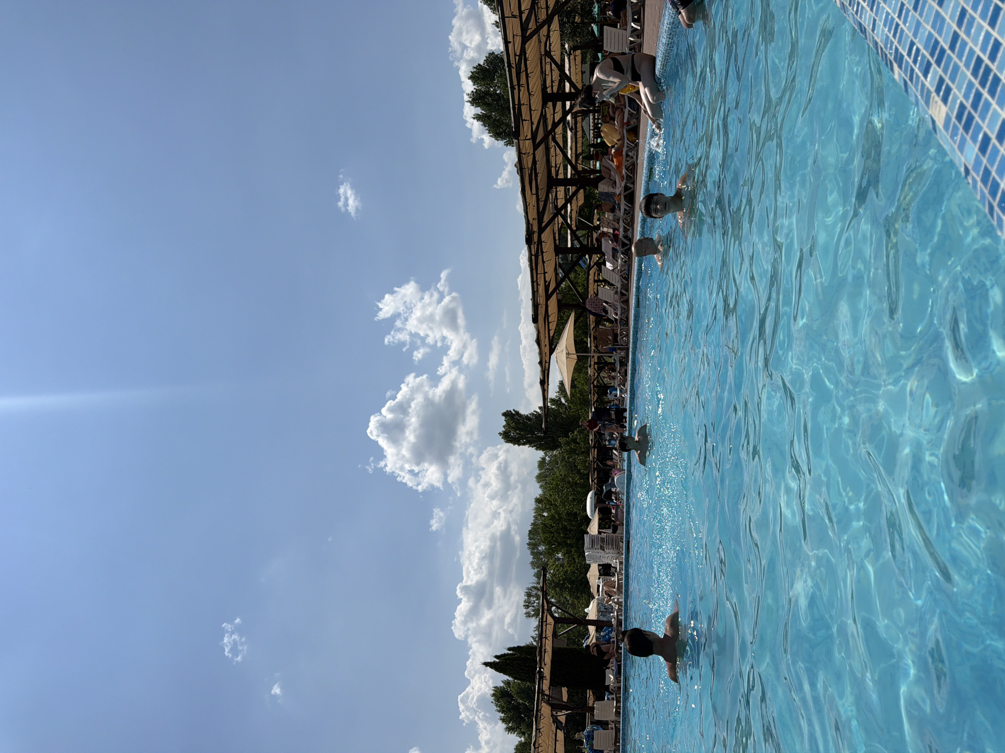 A high-angle, wide shot captures a bright, sunny day at a crowded outdoor swimming pool, likely located in or near the city of Sociteni, Moldova. The foreground is dominated by the crystal-clear blue water of the pool, with gentle ripples and sun reflections. Several individuals are visible in the water; in the immediate foreground to the left, a person with dark hair is partially submerged, facing away from the camera. To the right, another person with dark hair is in the water, looking towards the camera. Further back, more people are enjoying the pool.
The poolside is lined with lounge chairs and umbrellas, many of which are occupied by people sunbathing or relaxing. A wooden canopy structure with thatched roofing extends along the back of the pool area, providing shade. Beyond the pool area, lush green trees form a dense backdrop, suggesting a natural or park-like setting. The sky is a vibrant blue, punctuated by scattered white clouds, and a strong beam of sunlight angles down from the upper right corner of the frame. The overall scene depicts a lively and recreational atmosphere on a warm summer day.