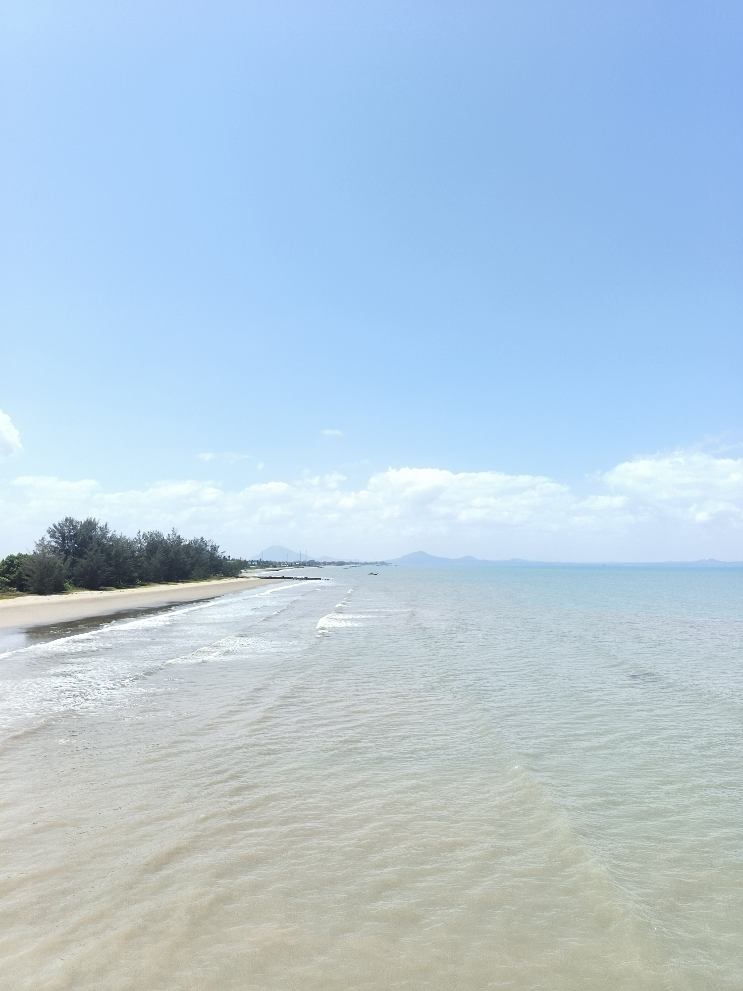 This image depicts a serene coastal landscape at Beringin Beach, located in the city of Beringin, Indonesia. The scene is dominated by the vast expanse of the ocean and a clear blue sky with scattered white clouds. The foreground features the gentle lapping of waves on a sandy shore, with the water appearing calm and shallow, showing subtle ripples and reflections.
The shoreline is lined with a dense growth of dark green trees, characteristic of a tropical environment, which creates a stark contrast with the lighter hues of the sand and sea. In the distance, the horizon is marked by a faint line of mountains and some man-made structures, suggesting the presence of a settlement or a port. A few small boats are visible on the water, indicating some maritime activity, though the overall impression is one of tranquility and natural beauty. The lighting suggests it is daytime, likely late morning or early afternoon, with soft sunlight illuminating the scene.