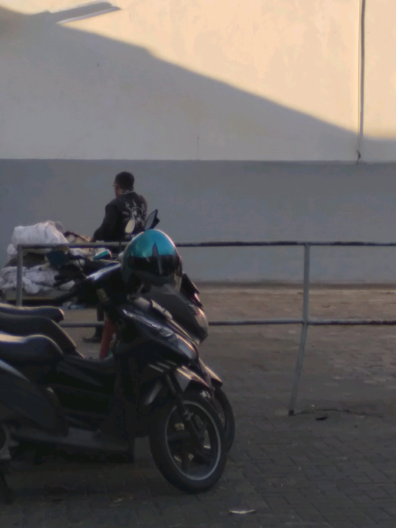 In Cimahi, Indonesia, a person wearing a black jacket with a graphic design is seated on a motorcycle. Another motorcycle, with a teal helmet on its seat, is parked in the foreground. A metal railing and a pile of white fabric are visible behind the motorcycles. The background features a light-colored wall with a large shadow cast upon it. The ground is paved with dark square stones.