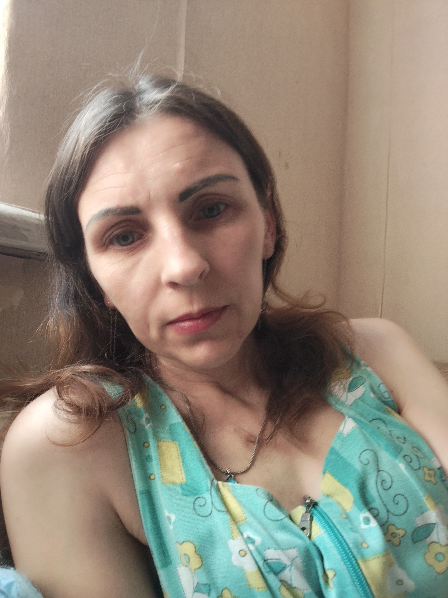 A woman with brown hair, blue eyes, and light skin is pictured from the chest up. She is wearing a turquoise and yellow floral patterned top with a zipper closure. She has a silver necklace and dangly earrings. The background is a light brown wall. The image is taken indoors. There is no discernible location in the city of Vinnytsia, Ukraine.