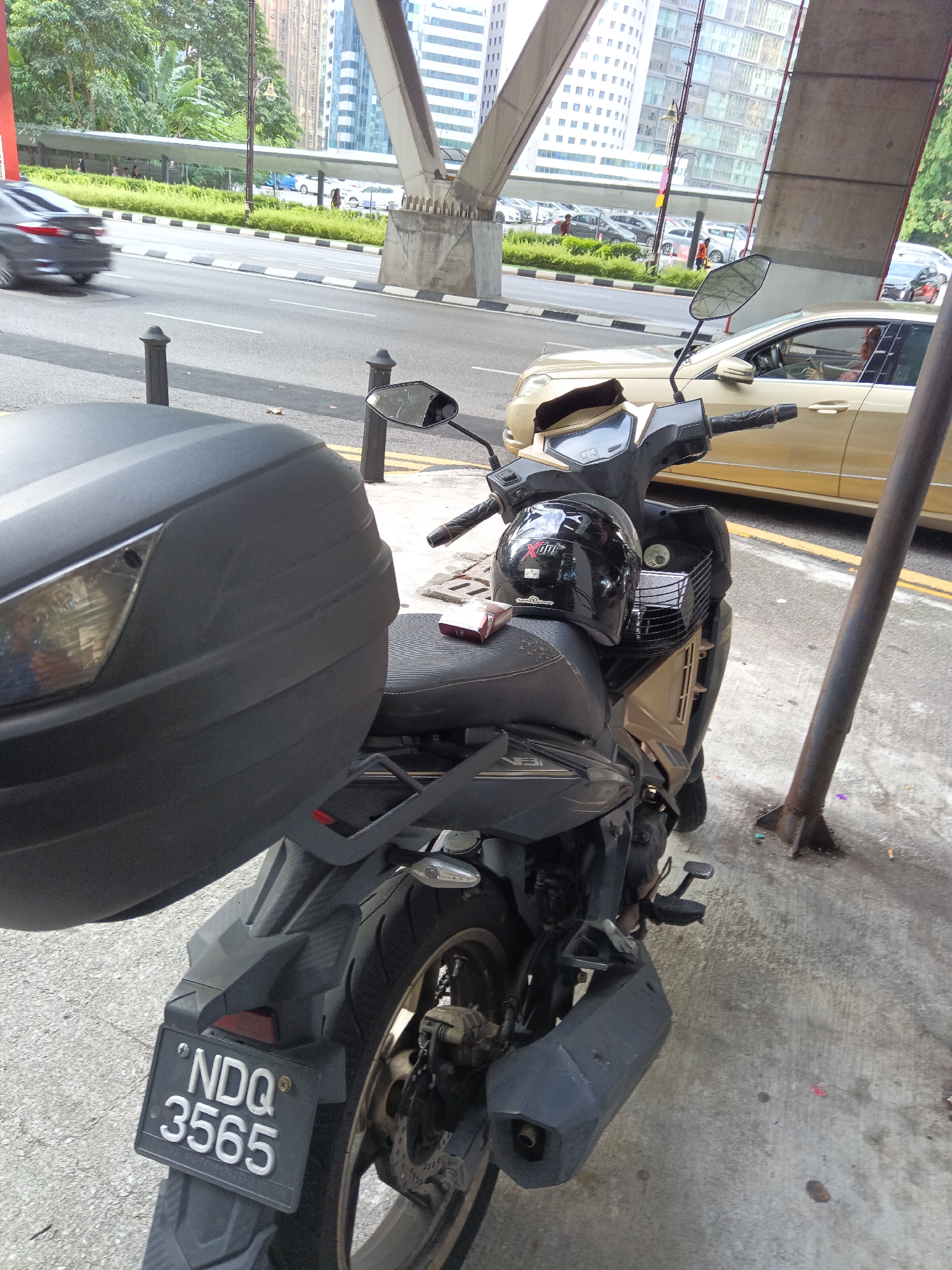 The image shows a street scene in Kuala Lumpur, Malaysia, featuring a parked motorcycle. The motorcycle, a dark color with gold accents, is positioned on a sidewalk next to a busy road. A black helmet sits on the motorcycle's seat, and a large black storage box is attached to the rear. The license plate of the motorcycle reads "NDQ 3565". In the background, traffic is visible on the road, including a gold sedan and other vehicles. Elevated structures, likely part of an overpass or bridge, dominate the upper portion of the frame. The environment appears to be urban, with buildings and greenery also present. The perspective is from a low angle, focusing on the motorcycle in the foreground.