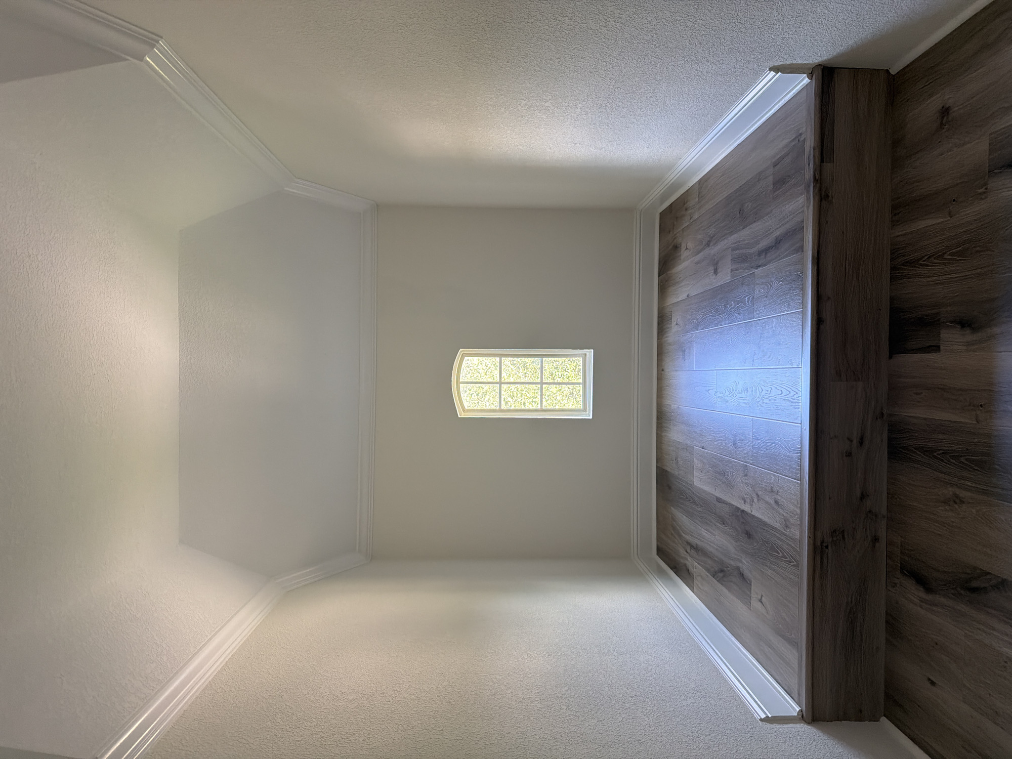 The image shows an empty, finished room with a raised platform floor covered in wood-look laminate flooring. The walls are painted a light cream color with textured wallpaper, and there is white crown molding along the top where the walls meet the sloped ceiling. A small, arched window with multiple panes is centered on the back wall, admitting natural light. The floor on the lower level also features the same wood-look laminate flooring. The scene is a generic interior view, likely of a residential property, but no specific Houston location is identifiable.