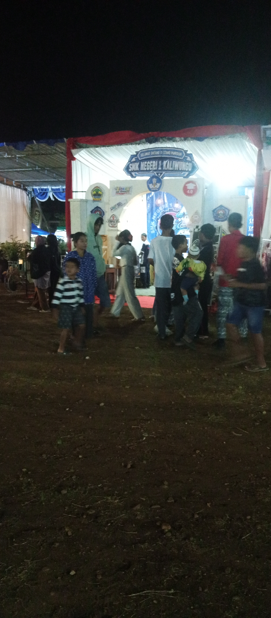 A group of people, mostly children and young adults, are gathered outdoors at night in what appears to be an event or fair in Kaliwungu, Indonesia. A large banner hangs above an entrance, reading "SELAMAT DATANG DI STAND PAMERAN SMK NEGERI 1 KALIWUNGU," which translates to "WELCOME TO THE EXHIBITION STAND OF SMK NEGERI 1 KALIWUNGU." The stand itself is brightly lit, with white tents and red draping, suggesting a formal display or event setup. Several individuals are walking through the area, some are standing and talking, and a few are holding or interacting with children. The ground is rough dirt, with some scattered debris and grass. The overall impression is a lively evening event, possibly an open house or exhibition hosted by the vocational school.