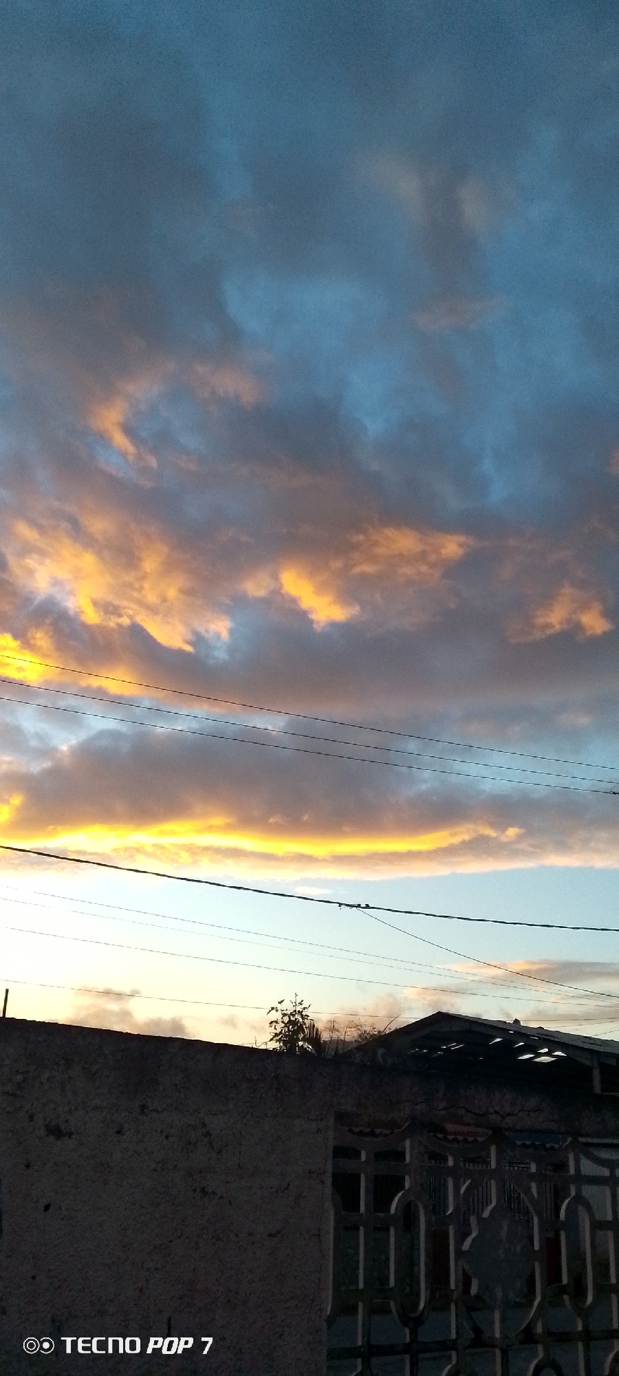The image captures a twilight scene, possibly sunrise or sunset, in Altagracia, Venezuela. The sky is predominantly filled with dark, greyish-blue clouds, some of which are illuminated with orange and yellow hues, indicating the presence of the sun below the horizon. Electrical wires are visible horizontally across the middle of the frame. Below the wires, a partial view of a building structure is visible. The building consists of a concrete wall and an ornate metal gate. A tree/bush is behind the wall. The bottom left corner of the image includes the imprint of a "TECNOP POP 7".
