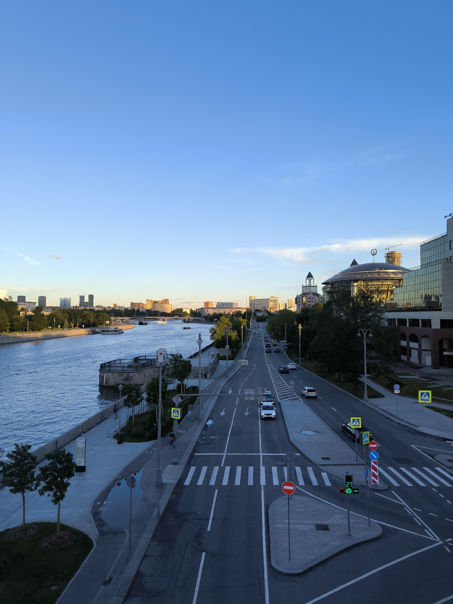 This is a high-angle, eye-level, full, vertical, outdoor, daytime, wide shot of a city riverfront and street. The sky is a clear, pale blue with a few wispy clouds. On the left, a wide, calm river with blue water occupies the left side of the frame. Along the riverbank is a paved walkway lined with trees and railings. A modern building with a glass facade and a dome-shaped roof is visible on the right side of the image, with other city buildings and skyscrapers in the background. A multi-lane road runs parallel to the river, with several cars driving on it. Pedestrian crossings and traffic signals are present on the road. The overall scene suggests a well-developed urban environment during daylight hours, likely in the late afternoon or early evening due to the warm lighting on the buildings.