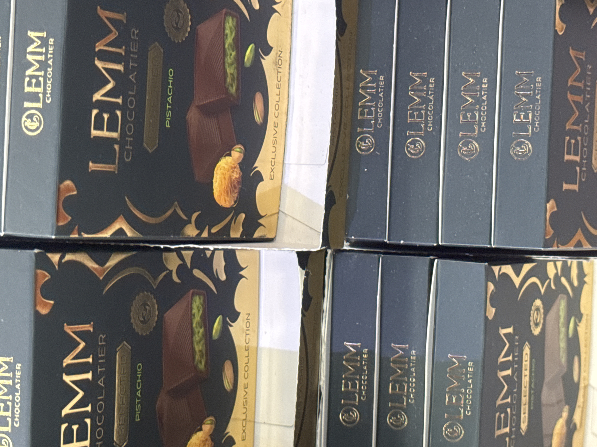 The image shows several boxes of "LEMM CHOCOLATIER" brand pistachio chocolates, stacked on top of each other. The boxes have a dark blue and gold color scheme with ornate patterns. The upper boxes display images of chocolate bars filled with pistachios and whole pistachios. The lower boxes are mostly visible as solid dark blue with the brand name and "CHOCOLATIER" printed in gold. The scene appears to be an indoor retail display, possibly in a shop or market, given the stacking and lighting. While the image is a photograph, there is no explicit indication of the specific location within Cenang Beach, Malaysia, beyond it being a commercial setting. No people are visible in the image.