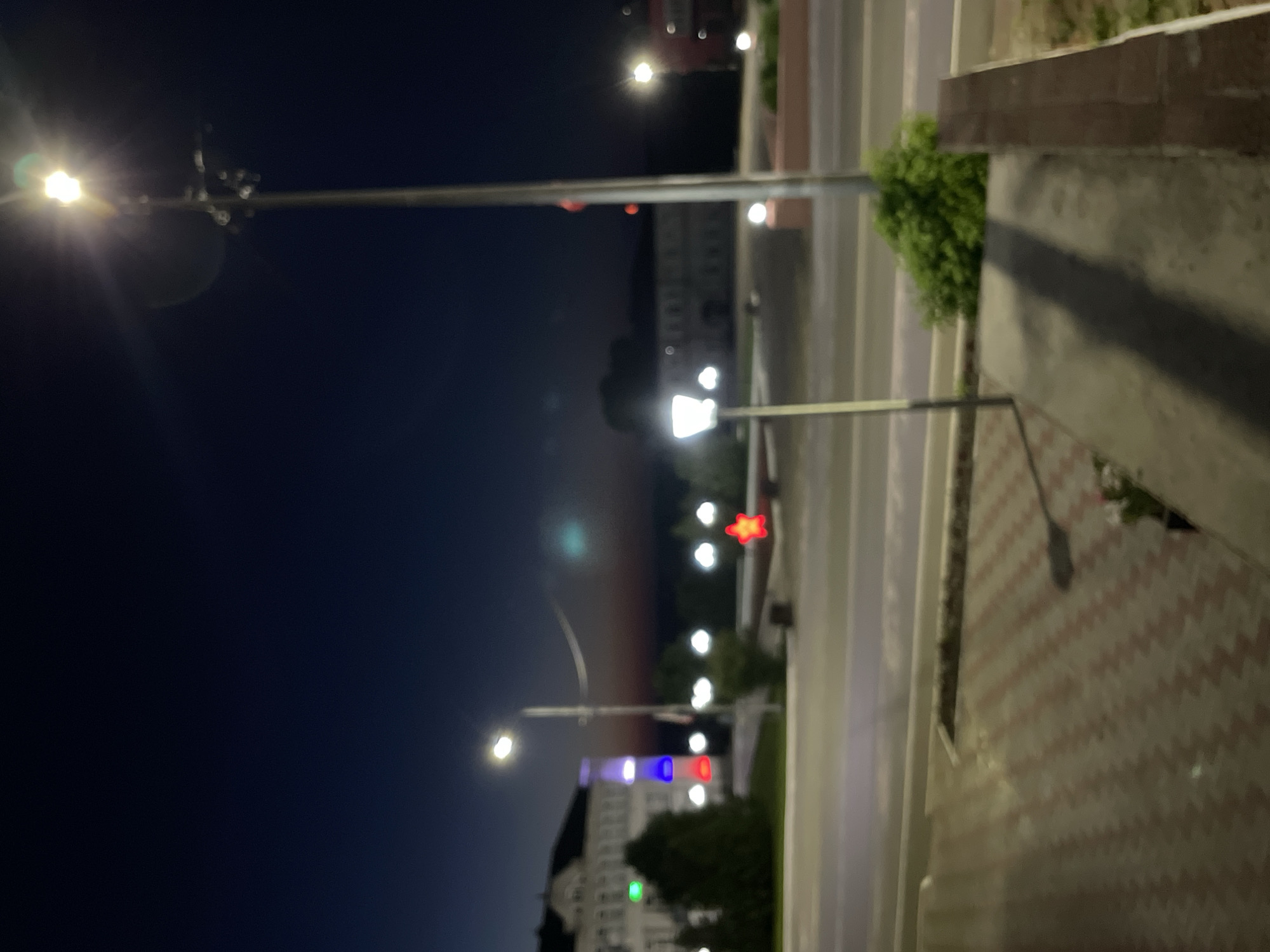 The image depicts a nighttime street scene in the city of Krasnye Baki, Russia. In the foreground, a patterned walkway of red and grey tiles leads towards a concrete surface with a dark shadow. To the right, a low brick wall is visible. In the middle ground, a street with two lanes is apparent, with a pedestrian crossing marked. A green bush is situated near the curb. Several streetlights are present, with one in the foreground emitting a bright light and causing a lens flare. In the background, buildings with illuminated windows line the street, and trees are scattered throughout the area. A prominent red star illuminated sign is visible in the distance. The sky is dark blue, transitioning to a lighter hue near the horizon, indicating twilight or a clear night.