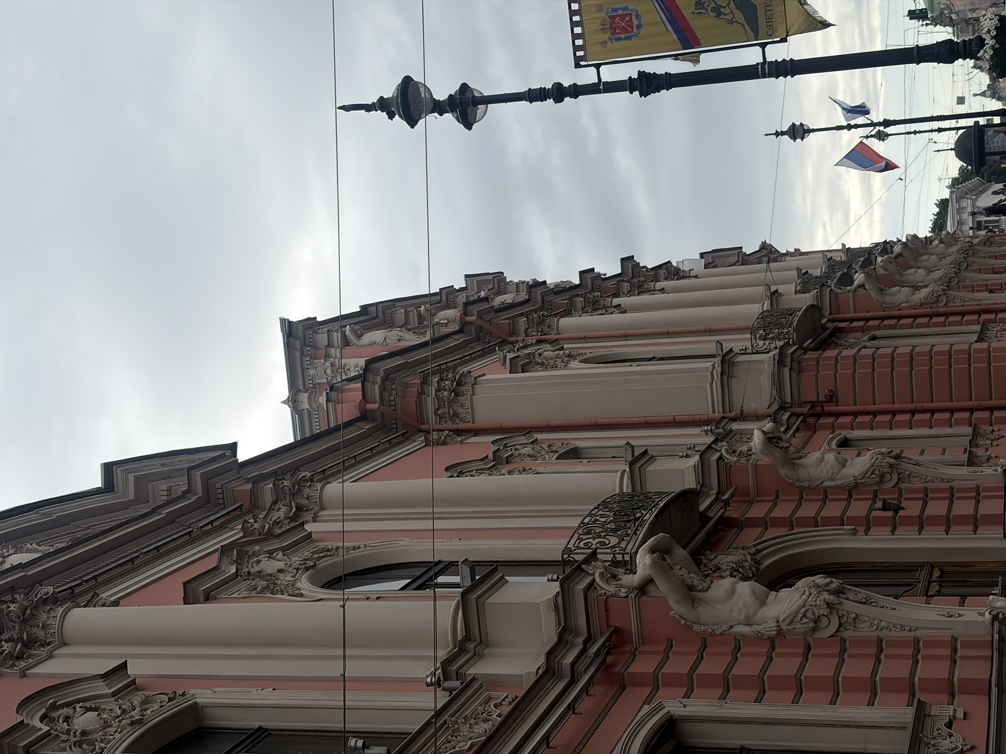 This is a street-level, eye-level, outdoor, slightly angled, vertical, medium shot of a building's facade in Saint Petersburg, Russia. The building is adorned with classical architectural elements, including columns and intricate carvings. Two prominent statues, appearing to be male figures supporting balconies, are visible on the lower facade. The building's exterior is painted a pale pink with cream-colored decorative elements. In the background, a tall, ornate street lamp stands, equipped with two lights, and a banner with a coat of arms and text is visible to the right of the lamp. Two small flags, one appearing to be the Russian flag, are attached to poles near the street lamp. The sky is overcast with a muted, cloudy appearance. Electrical wires are strung across the frame. The overall scene suggests an urban setting, likely a historic district, with a focus on the building's architectural details and surrounding street elements.