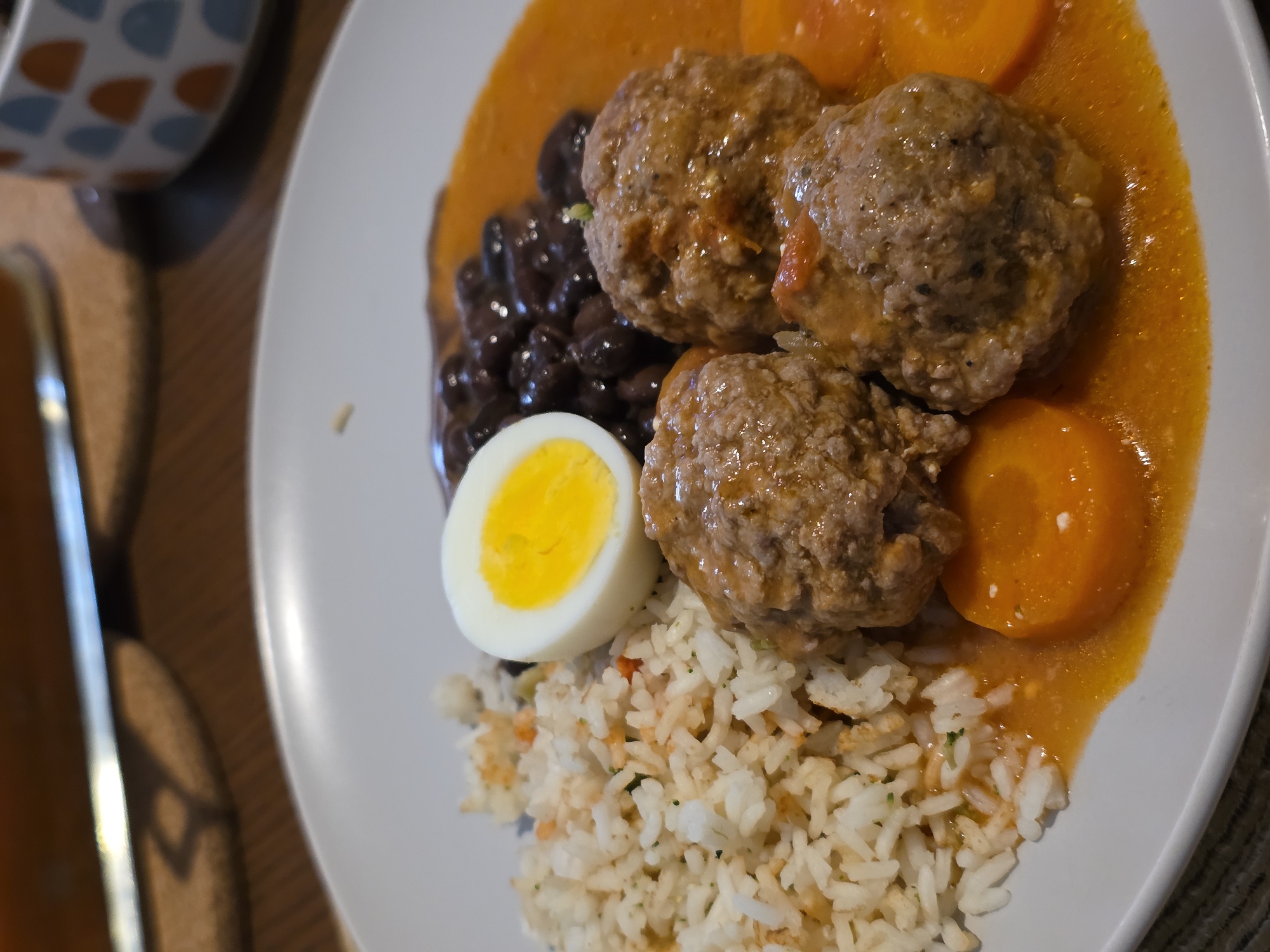 A top-down, medium shot depicts a white plate of food, including white rice with visible flecks of herbs and orange, three meatballs in a light orange sauce, black beans, half of a hard-boiled egg, and sliced carrots in the same light orange sauce as the meatballs. The food is arranged asymmetrically on the plate. Part of a cup with a blue and orange pattern and a piece of silverware are visible in the blurred background on a brown surface. The location in the city of Puebla, Mexico, is not determinable from the visual information.