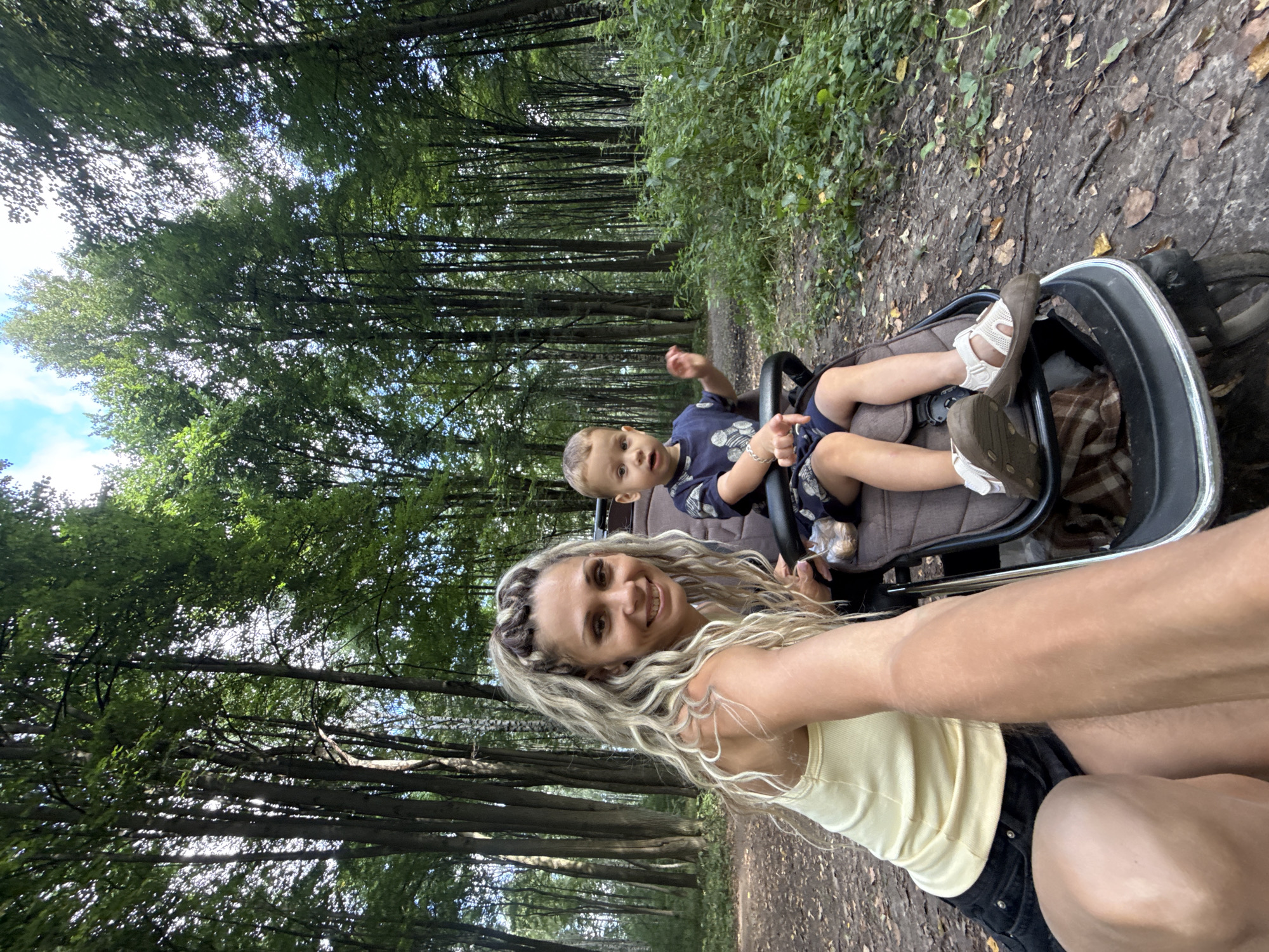 In Rusanovo, Russia, a woman with blonde, braided hair is taking a selfie in a forest with a young child in a stroller. The woman is wearing a yellow tank top and black shorts, and the child is wearing a dark blue outfit with a pattern. They are positioned on a dirt path with fallen leaves, surrounded by tall trees and lush green foliage. The sun is filtering through the leaves, creating dappled light. The woman is smiling at the camera, and the child appears to be looking in the same direction, with one hand raised.