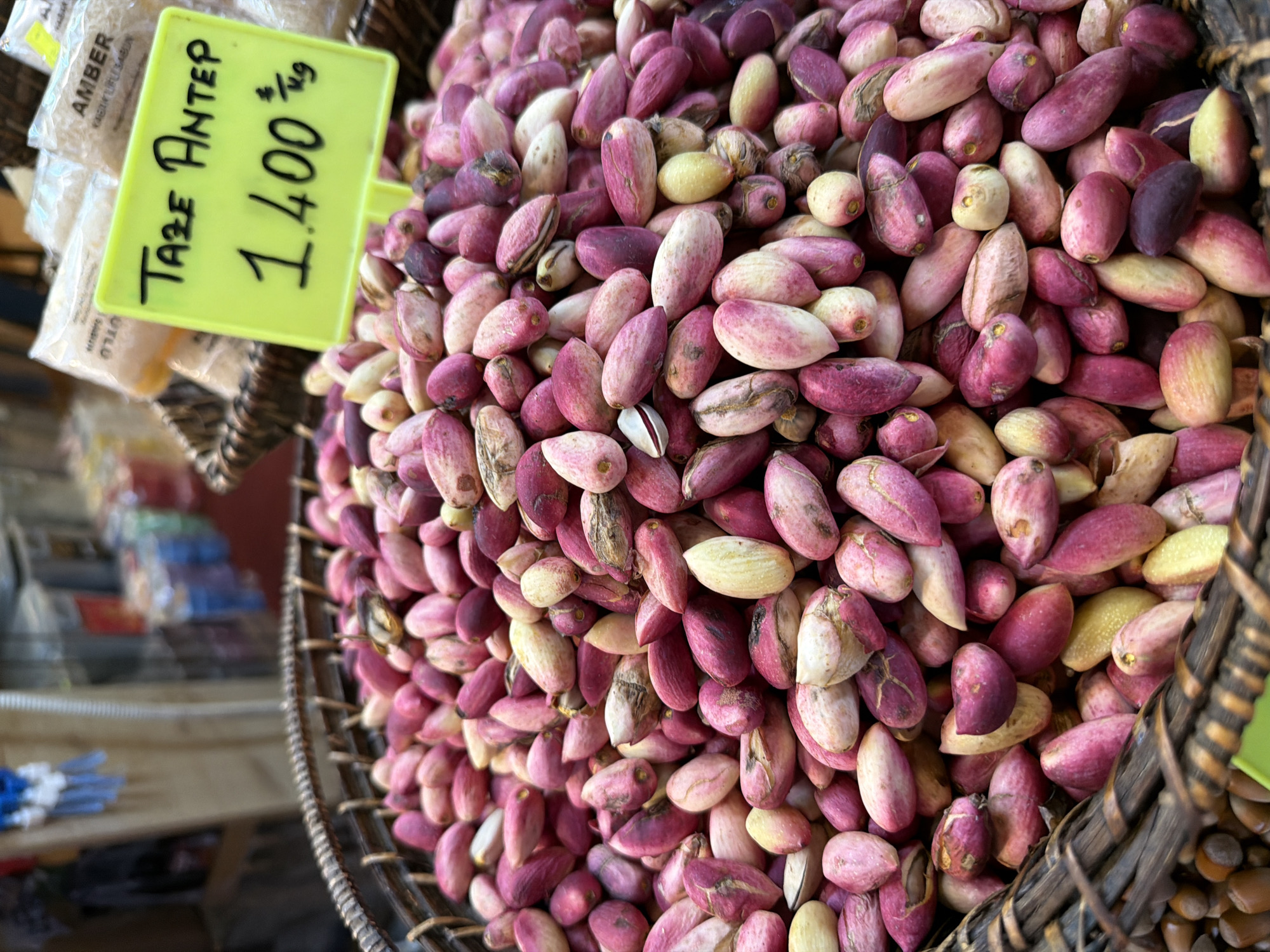 A close-up shot displays a woven basket overflowing with fresh pistachios. The pistachios vary in color, with shades of pink, purple, and a pale yellow. Some are partially shelled, revealing their vibrant interior. In the background, several clear plastic bags containing what appear to be dried goods are visible. A bright yellow price tag, handwritten in black marker, is partially obscured by the pistachios. The text on the tag reads "Taze Antep 1.400" with "kg" following the price, indicating that this is a market stall likely located in Kadıköy, Turkey, a district known for its vibrant markets. The overall impression is one of fresh produce for sale, likely in an outdoor or indoor market setting.