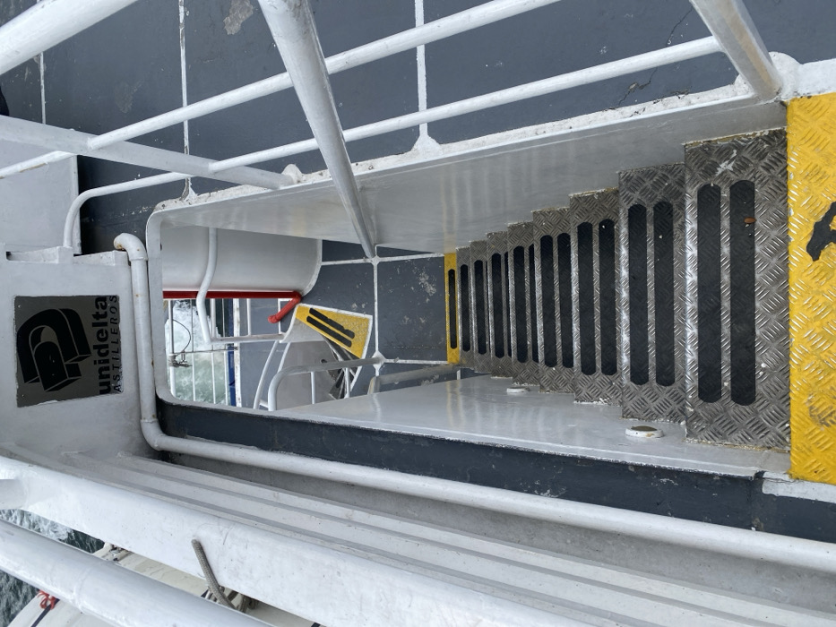A view looking down a metal staircase on a ship, likely a ferry. The stairs are gray with black stripes and yellow safety markings. A white railing surrounds the staircase, and the ship's metal deck is visible. The railing curves around a white box with the "unidelta ASTILLEROS" logo. The background shows glimpses of the sea and another part of the vessel. The overall impression is one of an industrial, functional space aboard a ship.