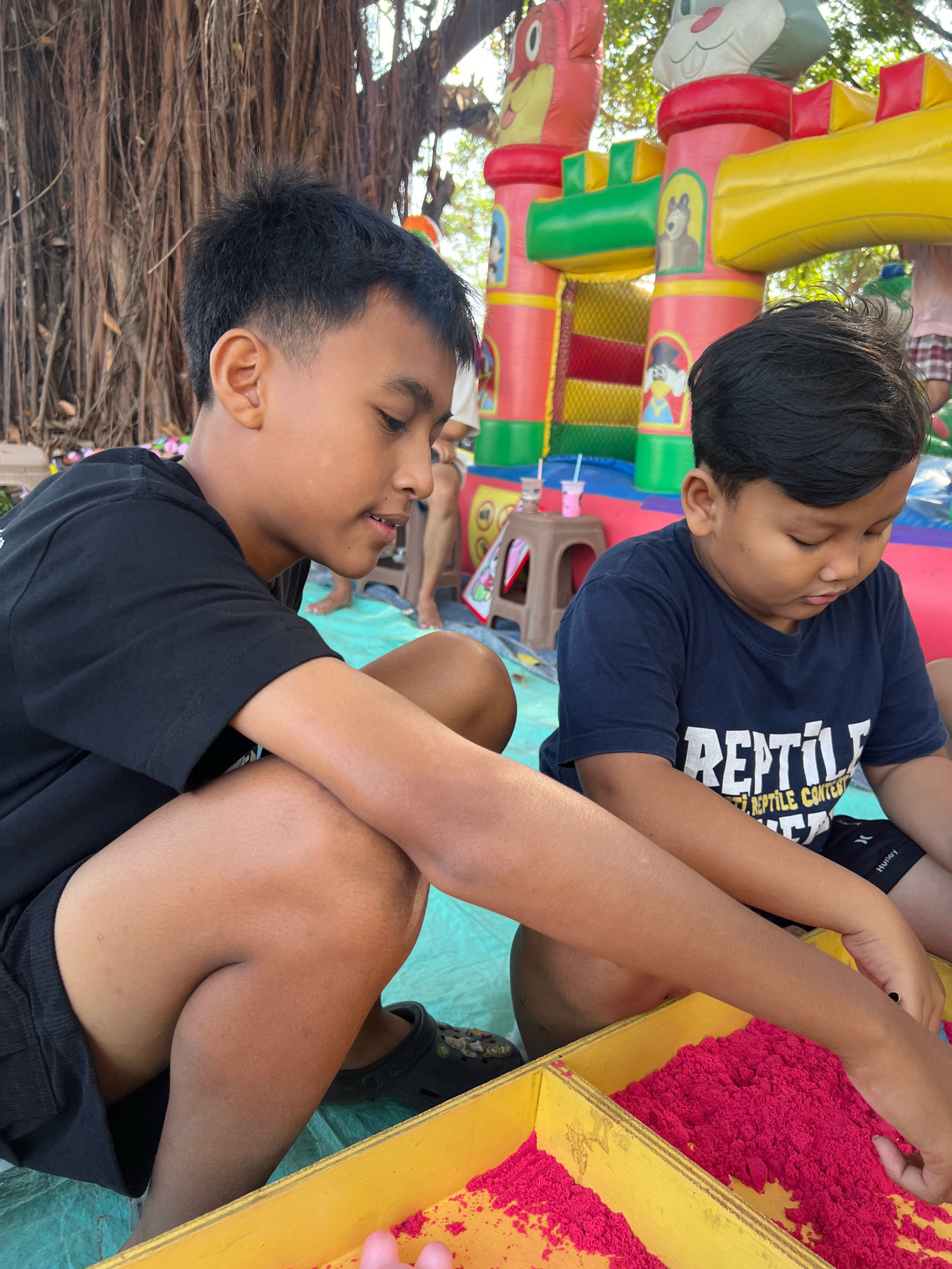 Two boys are crouching in front of a yellow wooden box filled with bright pink sand. The boy on the left, wearing a black t-shirt and black shorts, is looking down and to the right, talking. The boy on the right, wearing a navy blue t-shirt with the word "REPTILE" printed in large white letters, is also looking down, focused on the sand. Behind them, an inflatable bouncy castle with cartoon characters is visible, and large tree roots dominate the background. The scene appears to be outdoors in a public area, likely a park or event space, in Kecamatan Pati, Indonesia, given the presence of the bouncy castle and what appears to be local foliage.