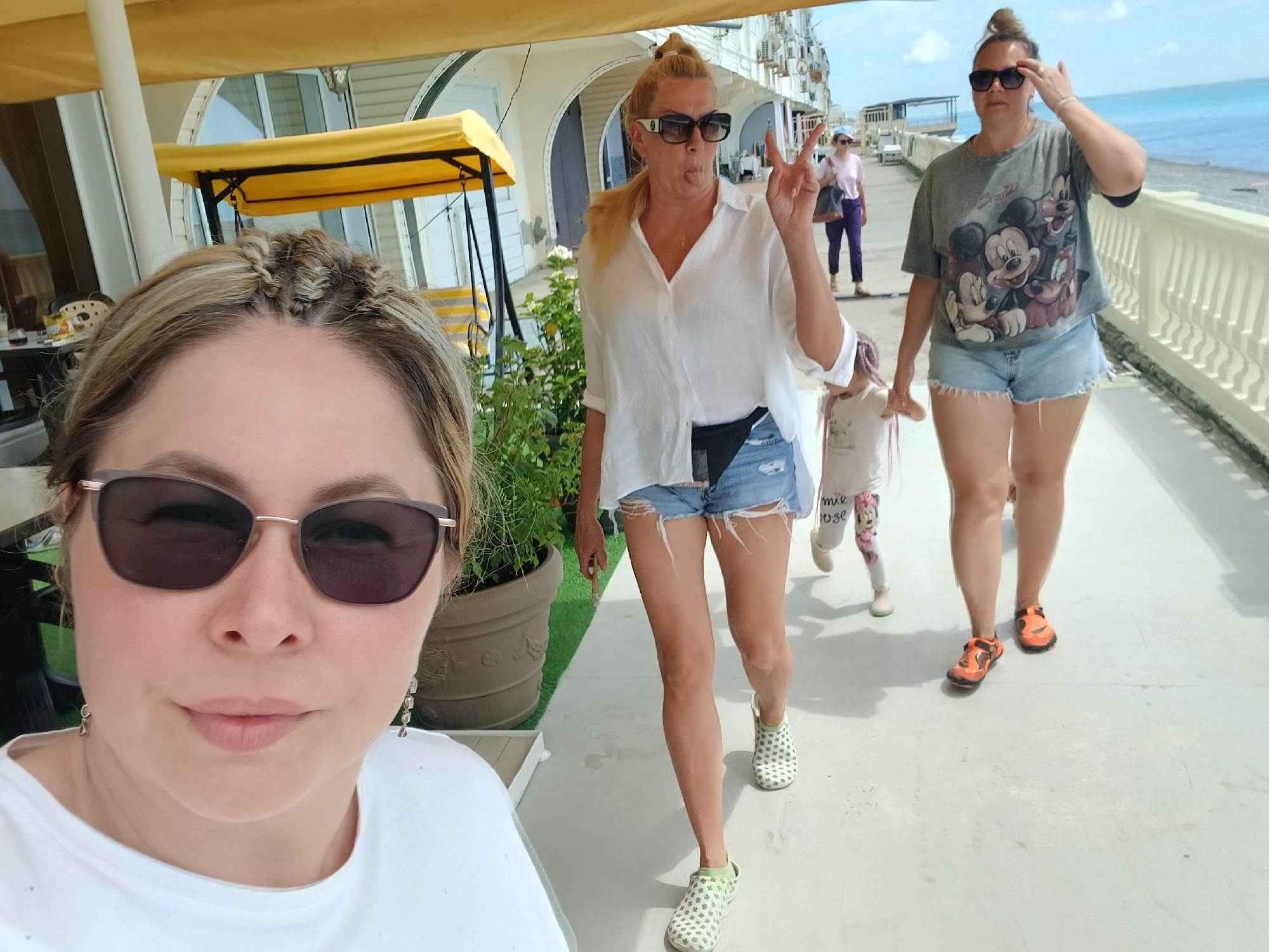 The image shows a group of people walking along a paved walkway next to the sea, likely in Sochi, Russia. In the foreground, a woman is taking a selfie, wearing sunglasses and a white t-shirt. To her right, another woman in a white shirt and denim shorts is sticking out her tongue and making a peace sign. Behind her, a young child in a white shirt with Mickey Mouse designs on the legs walks forward. Further back, a woman in a grey t-shirt with Mickey Mouse and denim shorts walks while touching her hair. The background shows a body of water, a railing, and buildings. A yellow canopy covers a seating area to the left. The overall scene depicts a casual outing in a sunny, outdoor environment.