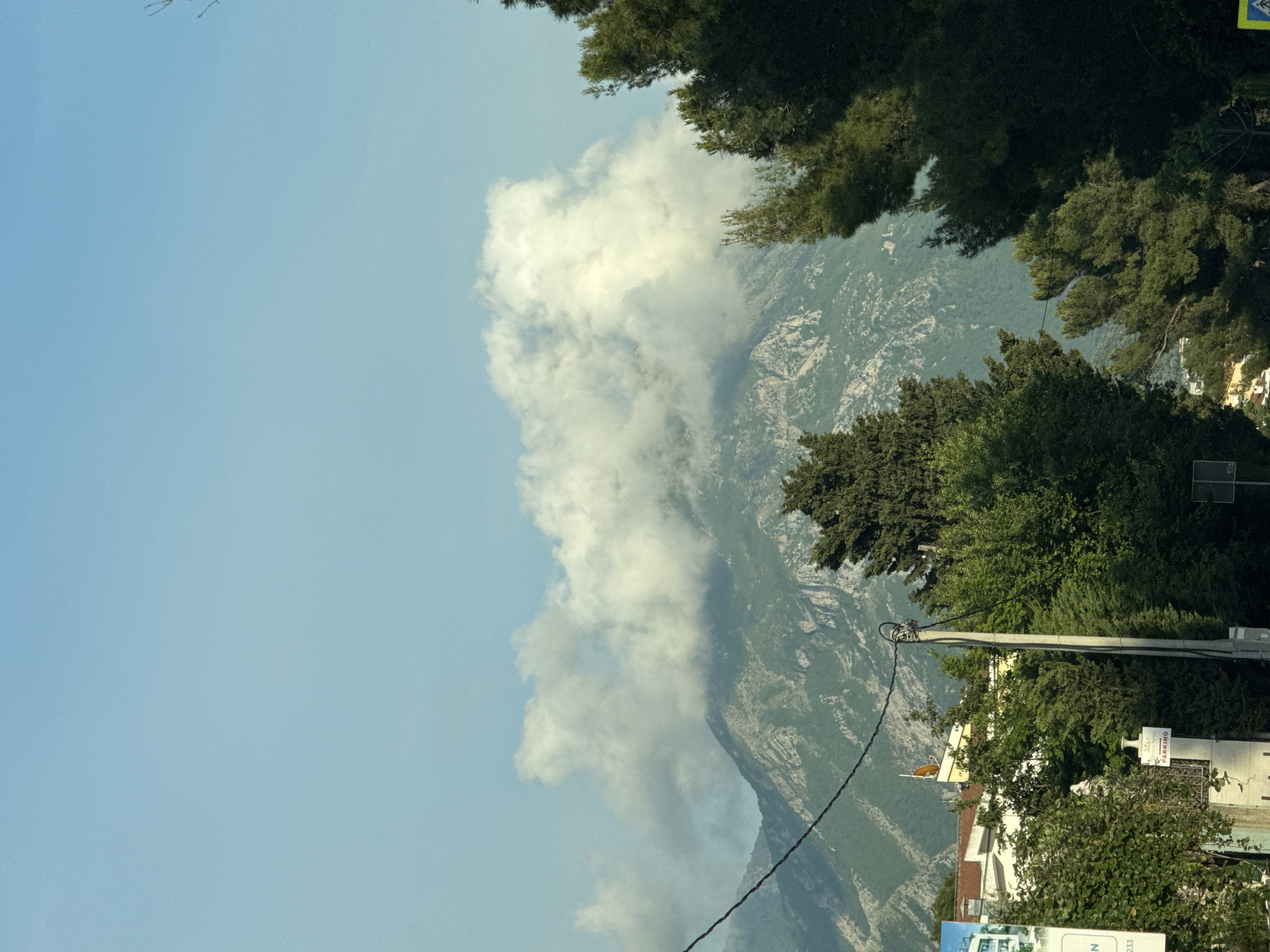 The image shows a landscape with a mountain range partially obscured by clouds. In the foreground, there are trees and glimpses of buildings, suggesting an urban environment at the foot of the mountains. A utility pole and power lines are visible, further indicating a populated area. The sky is blue, with varying cloud cover ranging from dense to sparse. Based on the mountainous terrain and the presence of urban elements, it is plausible that the location is Bar, Montenegro.