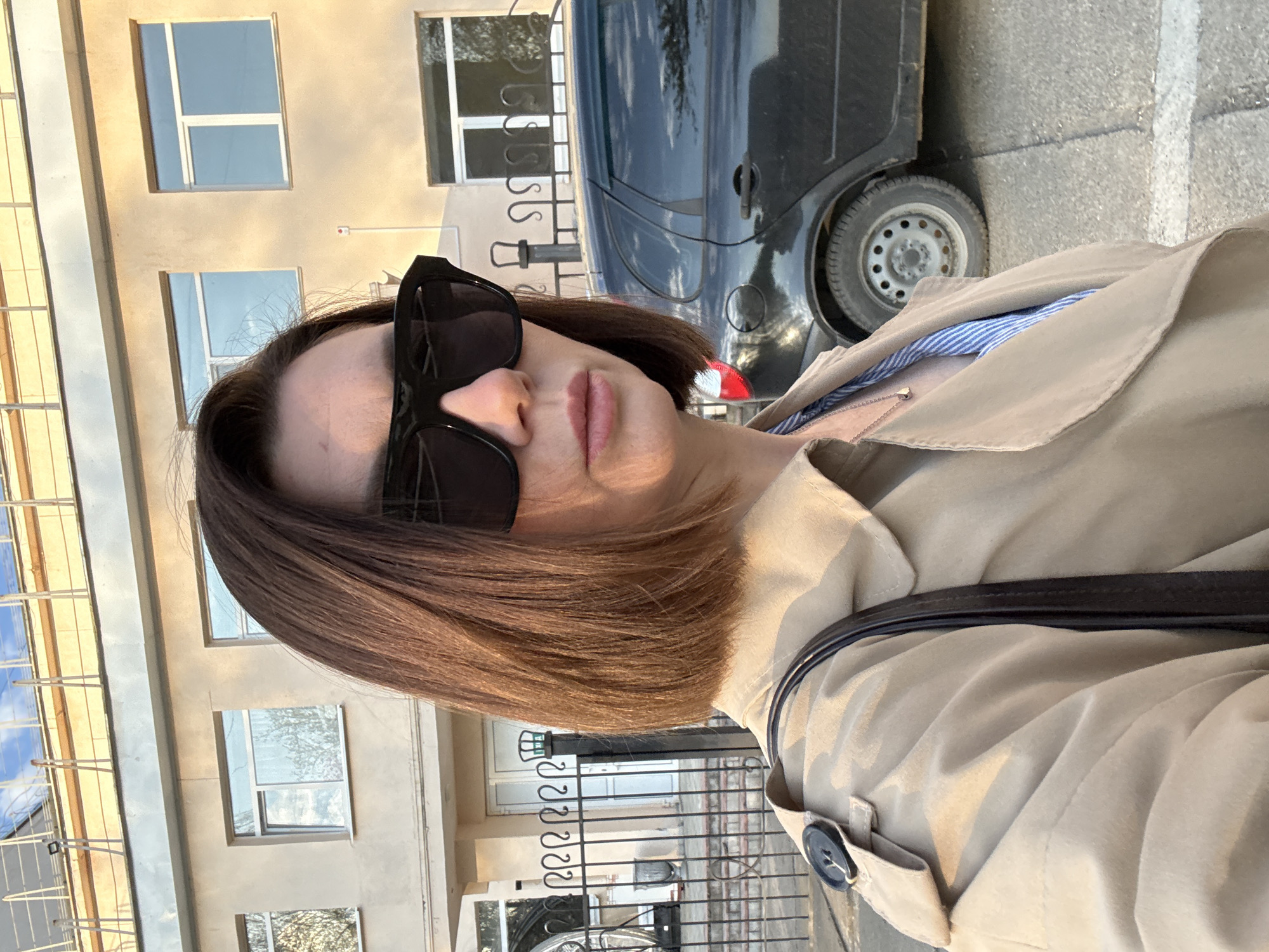 A woman is outdoors in Chaikovsky, Russia, based on identifying elements. She's wearing sunglasses and a beige trench coat. A dark-colored car is parked behind her on a paved surface with a white line. In the background, there's a multi-story building with rectangular windows and decorative metalwork. A metal fence with coiled designs is also visible. The image appears to be taken during daylight with noticeable shadows.