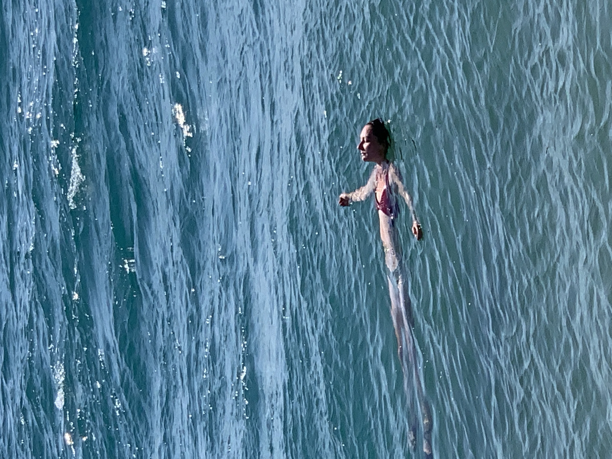 A person is floating on their back in the ocean. The person is wearing a red bikini. The water is a deep blue-green color and appears to be choppy. The sun is shining and reflecting off the water. The person's head is tilted back and their eyes are closed. Their arms are extended out to their sides. Their legs are also extended. The person appears to be relaxed and enjoying the water. The location is unknown.