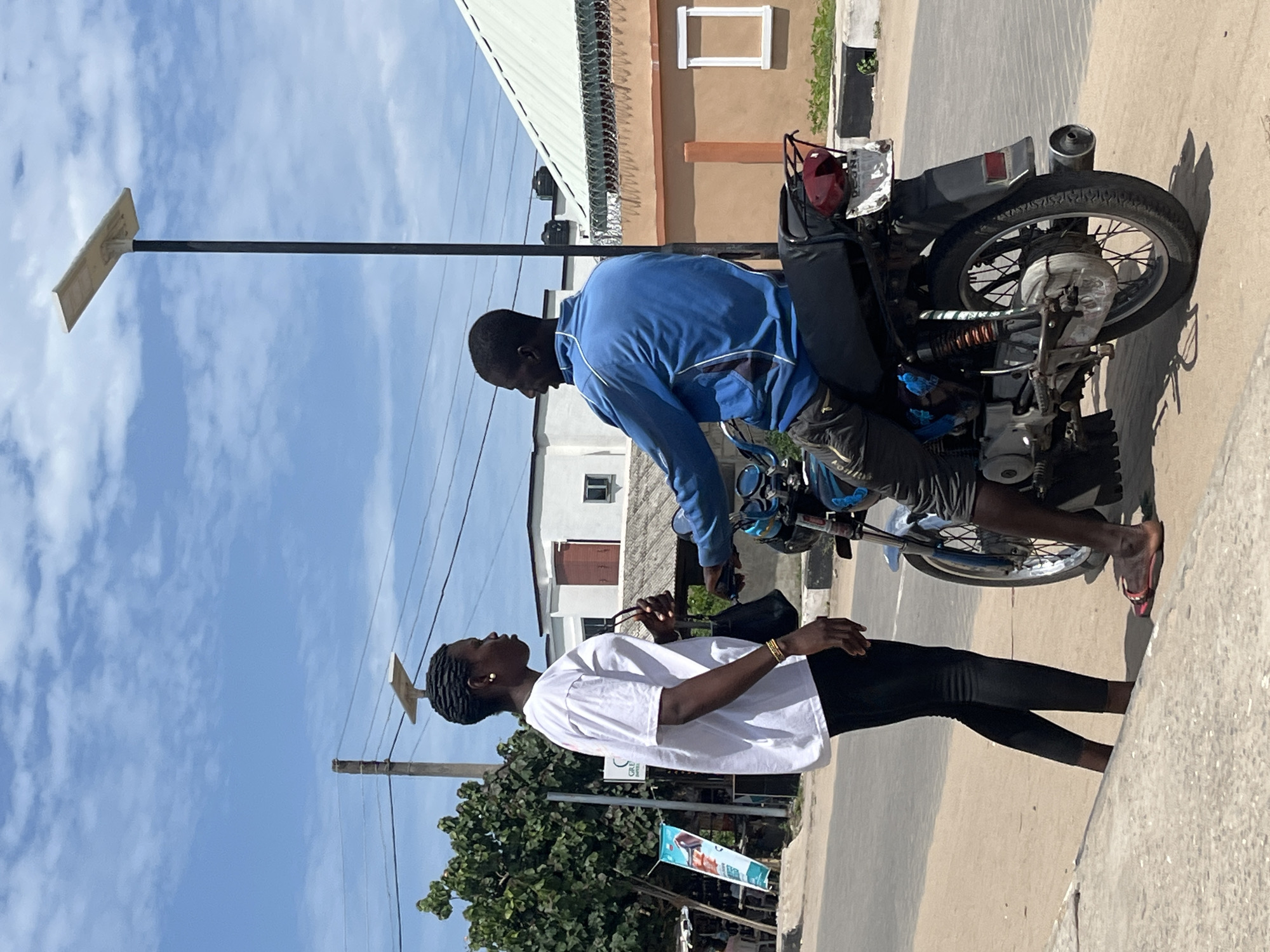 The image shows a man on a motorcycle interacting with a woman on a street in Badagry, Nigeria. The man is wearing a blue jacket and shorts and is sitting on the motorcycle. The woman is wearing a white t-shirt and black leggings. They are facing each other. A street light is visible in the background, along with buildings and trees. A banner advertisement is beside the road.