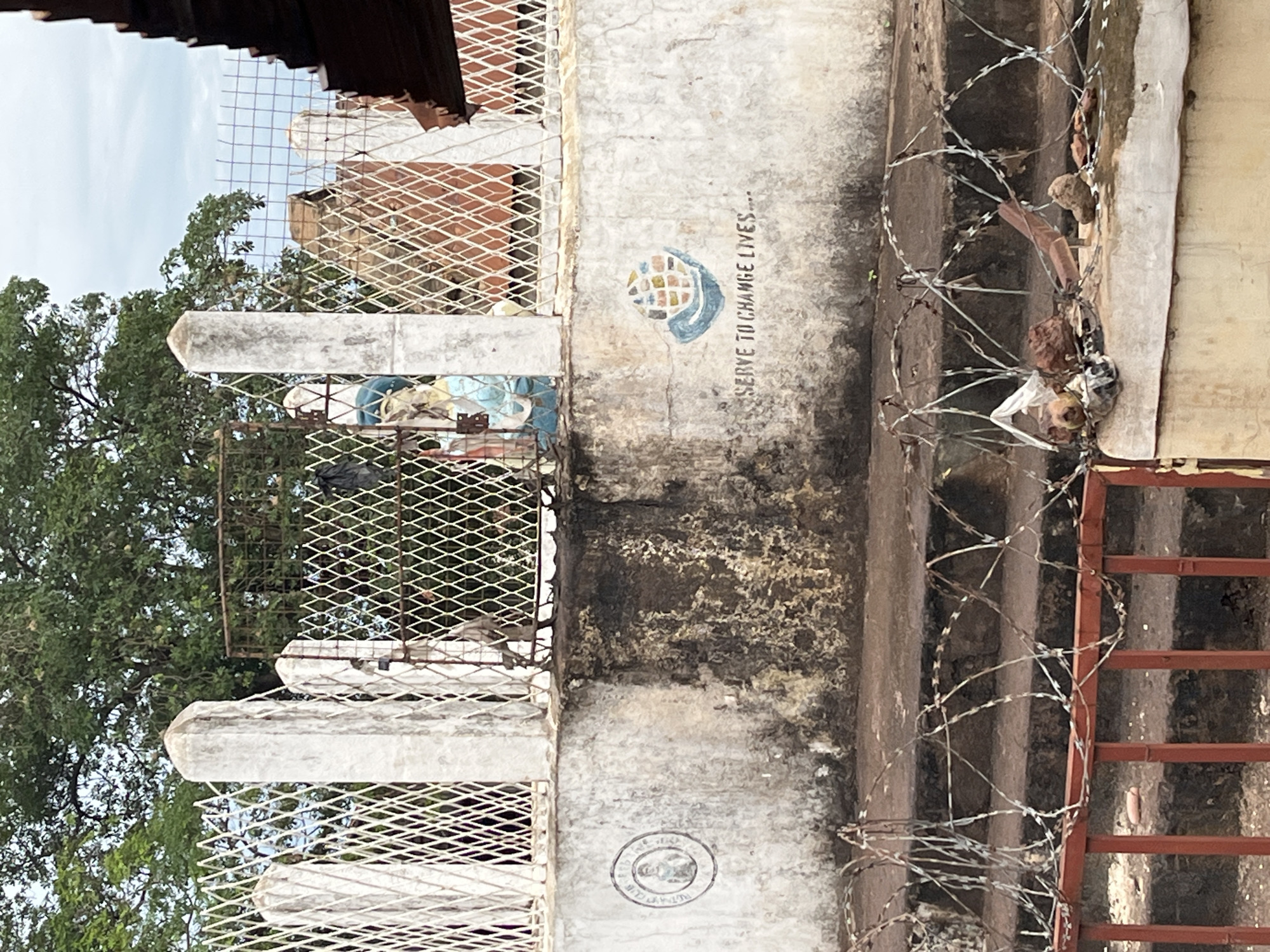 This image, captured in Idah, Nigeria, depicts a weathered outdoor scene centered on a concrete retaining wall topped by a metal fence. The wall, light-colored and stained, particularly with a large dark patch on its lower right section, supports a fence constructed from white concrete posts and white diamond-patterned metal mesh. A darker, possibly rusted, gate-like section is centrally located within the fence line. Behind the fence, a set of worn concrete stairs ascends.
Two distinct emblems are painted on the concrete wall: on the left, a faded circular logo contains a portrait of a person and the text "ROTARACT CLUB IDAH"; on the right, a stylized globe logo with multicolored squares and a blue wave-like base is accompanied by the motto "SERVE TO CHANGE LIVES....". Behind the fence, a light blue and white statue, possibly religious, is visible slightly to the left of the center, with another indistinct lighter-colored figure or object to its right.
In the foreground, a red metal barrier or gate is entangled with coils of razor wire (concertina wire). Various pieces of debris, including small rocks, plastic, and other refuse, are scattered on top of this barrier. The background features lush green trees under a partly cloudy sky. A dark-colored, possibly tiled or corrugated, roofline is partially visible on the far right edge of the frame. No individuals are actively present in the scene, though the emblems suggest human organizational activity.