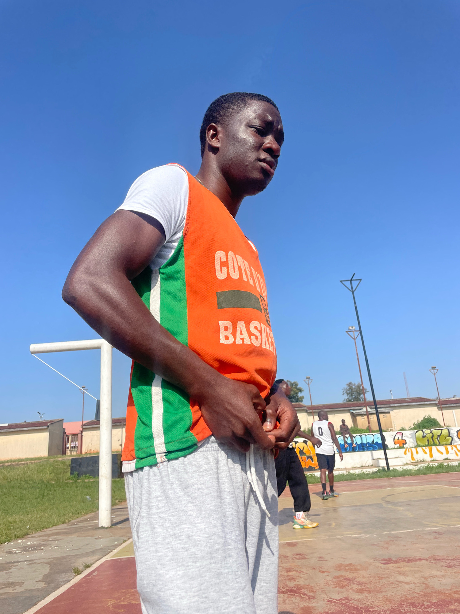 The image shows a young Black man standing on a basketball court on a sunny day. He is wearing a white t-shirt and an orange and green basketball jersey with the word "BASKET" printed on it. He is looking to his right with a serious expression. In the background, there are other people on the court and a building with graffiti on it. The basketball court appears to be in an urban area, possibly in the city of Jos, Nigeria, given the context. The sky is clear and blue.