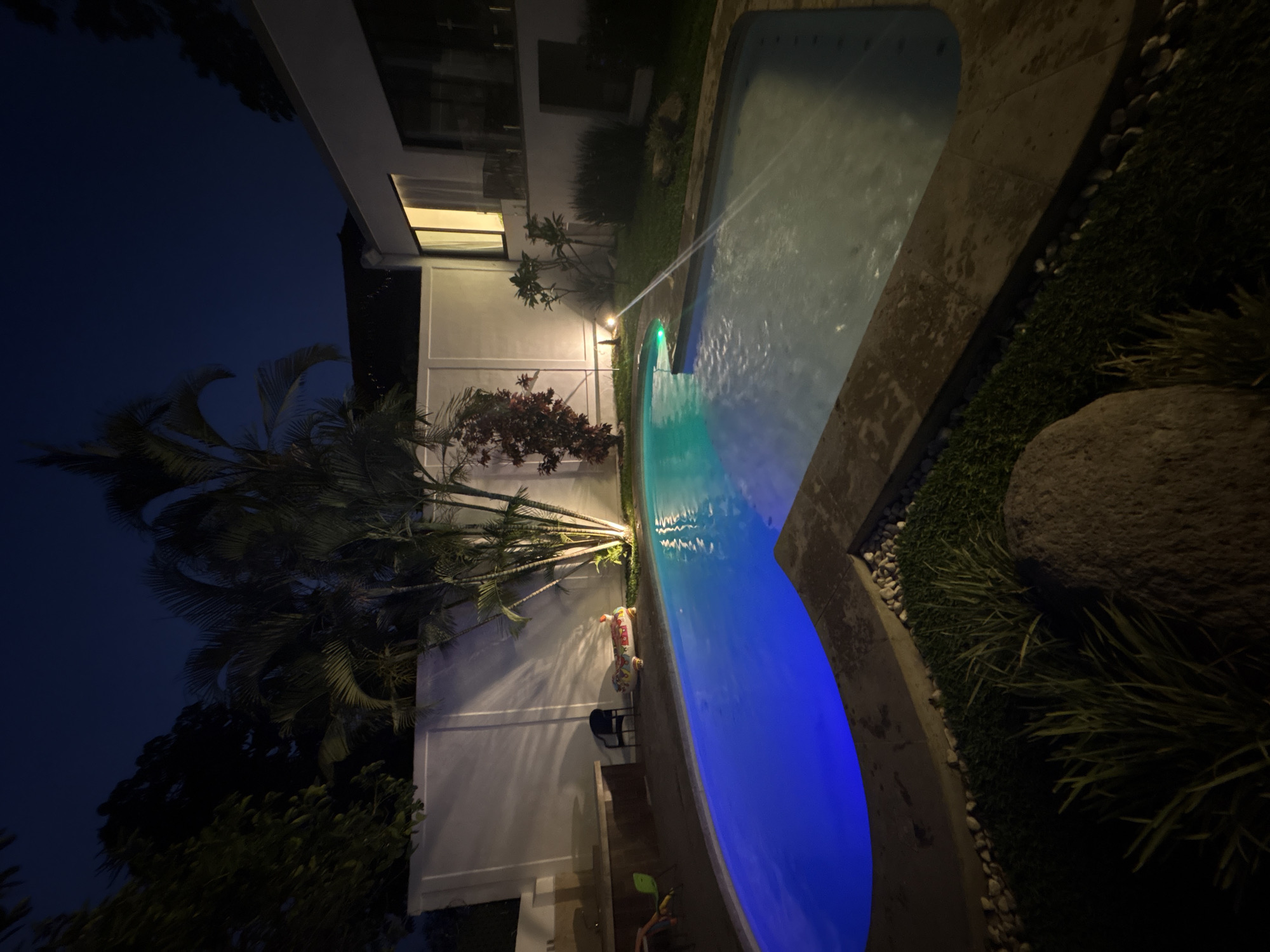 At Tres de Mayo, Mexico, the scene depicts an illuminated outdoor area at night, featuring a swimming pool with multicolored lighting that transitions from blue to green and white. The pool has a water feature, possibly a fountain, spraying water into the pool.
Adjacent to the pool, there's a patio with an inflatable alligator, a black chair, and toys. The area is bordered by a white building with lit windows and a surrounding landscape of trees, bushes, and grass. The overall scene is a mix of a recreational area integrated with natural elements.