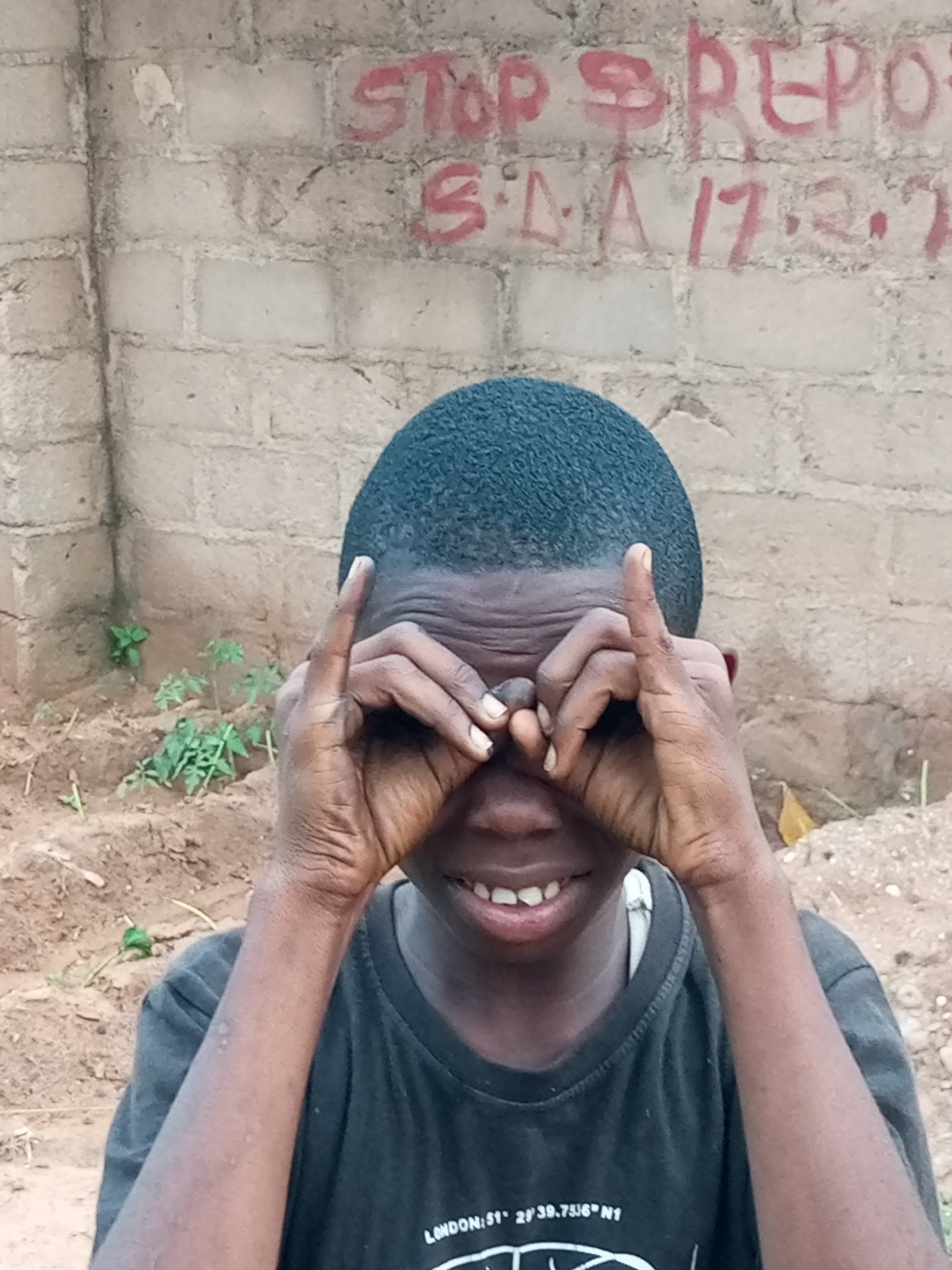 A young Black boy with short hair and wearing a black t-shirt smiles at the camera, holding his hands up to his eyes as if wearing binoculars. The t-shirt has white text on it that reads "LONDON: 51° 2' 39.7516" N1". The boy is positioned in front of a light brown brick wall with red graffiti. The graffiti says "STOP REPORT" on the top line, and "S.L.A. 17.2." on the second line. The ground in front of the wall is dirt with some small green plants. This scene is located in Tempure, Nigeria.