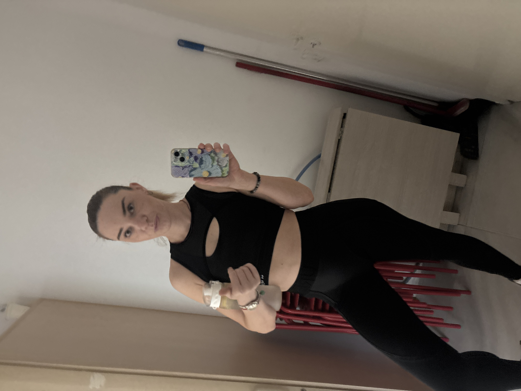 A woman in athletic wear is taking a mirror selfie. She is wearing a black sports bra with a cutout detail and black leggings. She is holding a smartphone with a colorful floral case in her left hand, and a dumbbell in her right hand. The dumbbell has a white cloth wrapped around its end. The woman has her hair pulled back and is looking at the camera with a neutral expression.
The setting appears to be an indoor space, possibly a gym or a room in a house. In the background, there are cleaning tools leaning against a wall, including a mop and a broom with red handles. There is also a light-colored cabinet or shelf unit. The lighting suggests it is daytime, but there are no clear indications of the time of day or weather conditions. The overall impression is that the woman is preparing for or is in the middle of a workout.