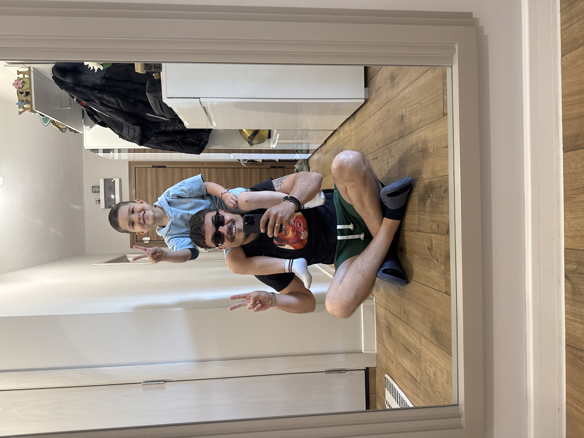 A mirror selfie shows a man sitting cross-legged on a wood floor, with a child on his shoulders. The man wears sunglasses, a t-shirt, and dark shorts, holding a phone up to take the picture, and giving a peace sign. The child wears a light blue shirt and also holds up a peace sign. They are indoors in an entryway with a closet. The location appears to be an apartment in an unknown city.