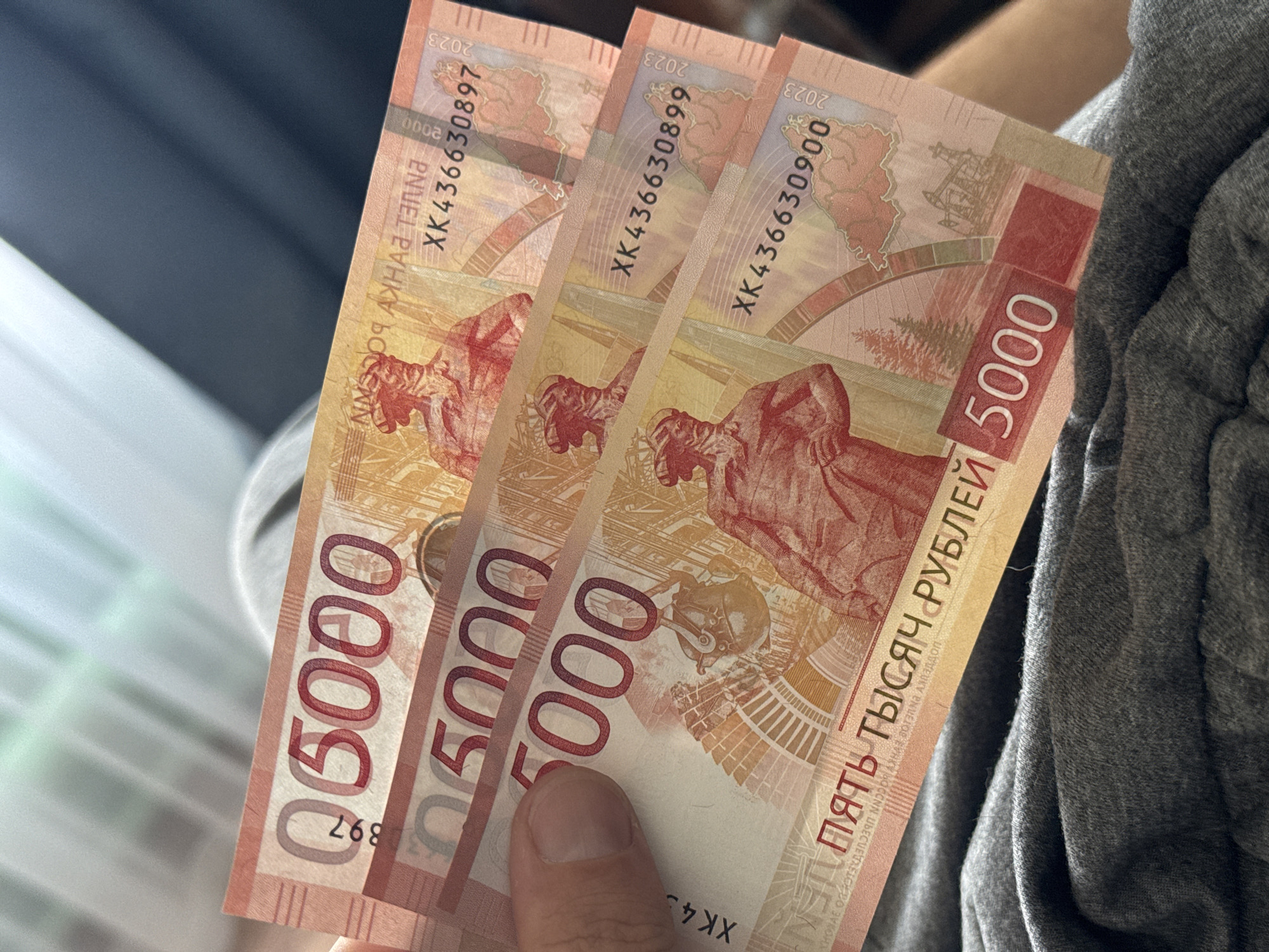 A person is holding three Russian banknotes worth 5000 rubles each. The banknotes feature images of the city of Khabarovsk, Russia, and its landmarks. The banknotes bear serial numbers XK436630897, XK436630899, and XK436630900. The year 2023 is printed on the banknotes. The person is wearing a gray garment.