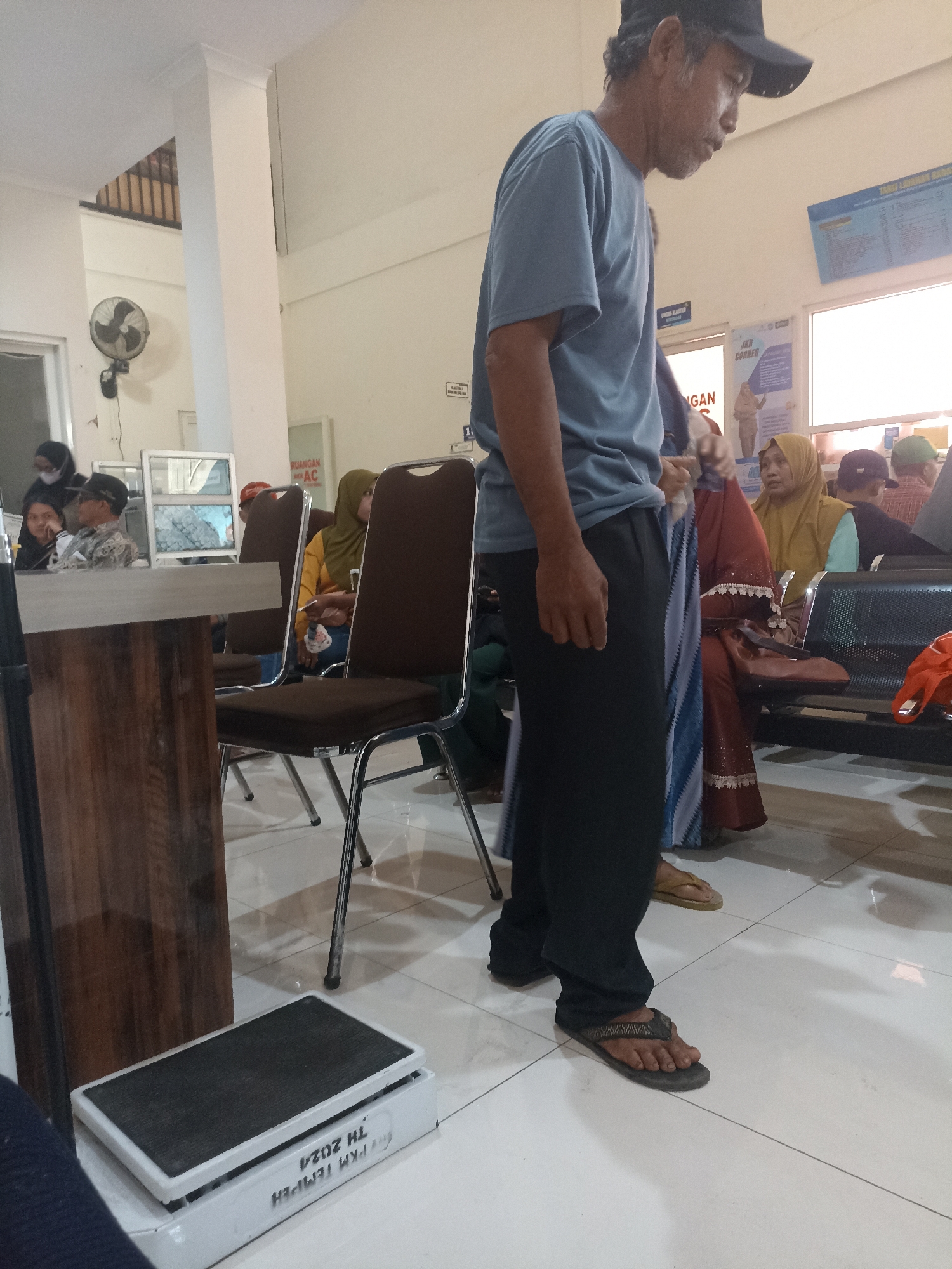 In a waiting area in Tempeh, Indonesia, a man wearing a blue t-shirt and dark pants stands near a digital scale. He is also wearing a black baseball cap and sandals. Other people are seated in the background, including a woman in a yellow hijab and a woman in a red and blue patterned dress. The room appears to be a public waiting area, possibly a clinic or government office, with chairs, a reception desk, and a wall-mounted fan visible. The lighting is bright, suggesting an indoor environment with artificial illumination. The floor is tiled and appears clean. A scale with Indonesian text is positioned on the floor near the foreground.
